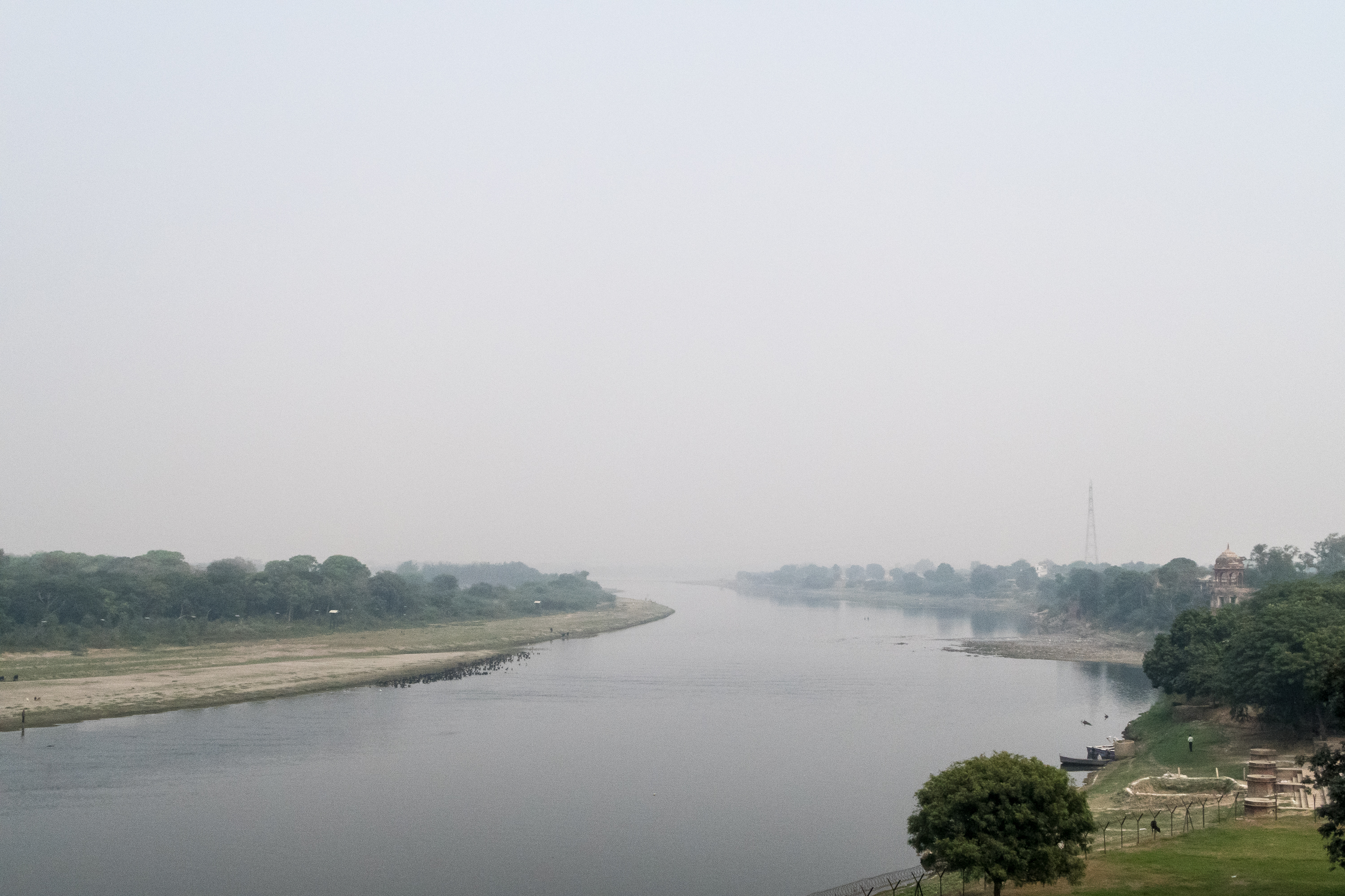 A serene river flows calmly under a hazy sky, bordered by green trees and vegetation. The foggy atmosphere softens the distant view, reminiscent of picturesque riverbanks in India. A pagoda and small buildings are visible on the right bank, exuding tranquility and natural beauty with minimal human activity.