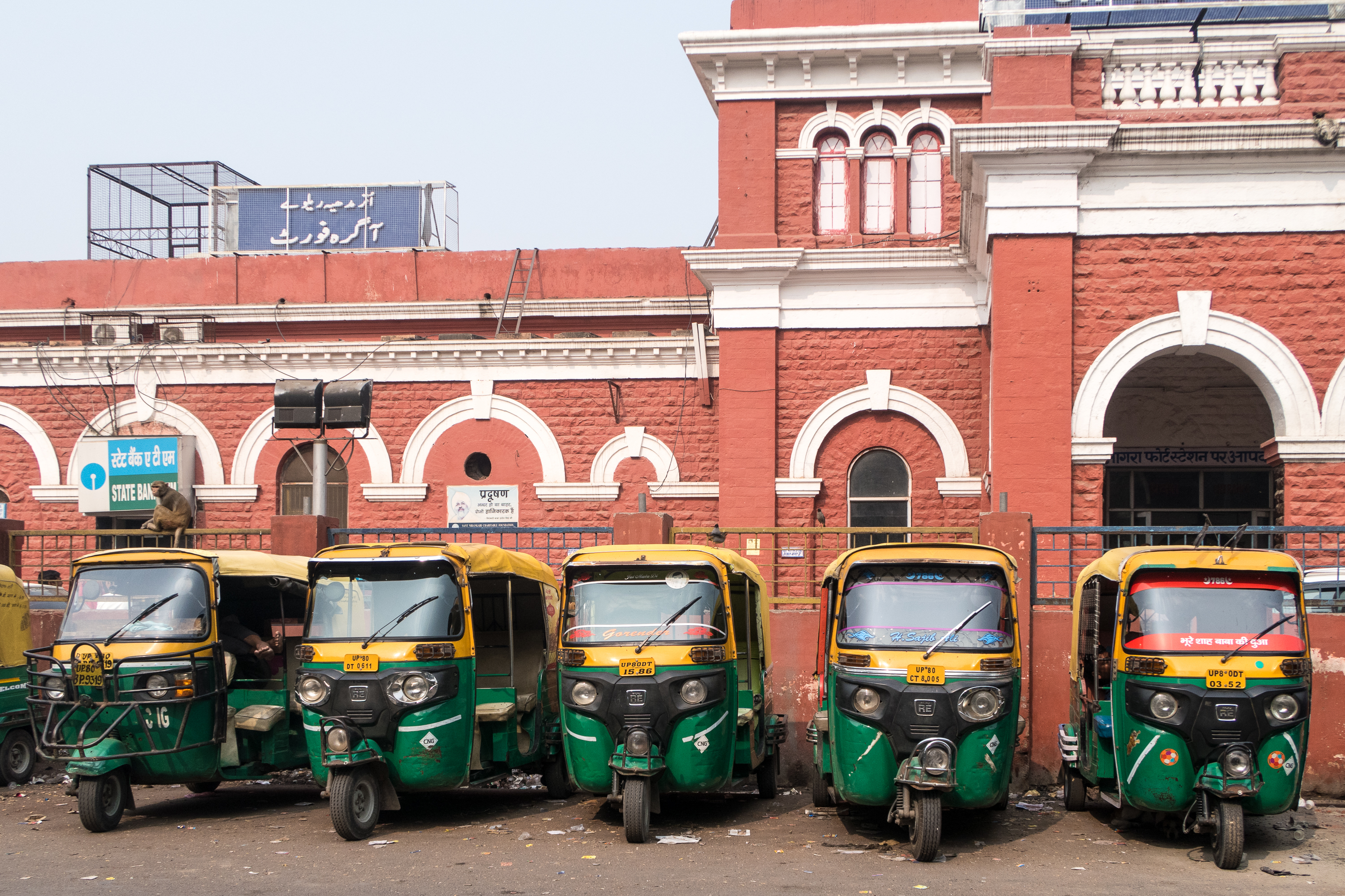 Six colorful auto rickshaws are parked in a row in front of a historic red brick building with ornate white trim, typical of India's rich architecture. The building has arched windows and a blue sign with text in an Asian script. A clutter of items and some debris are visible around the rickshaws.