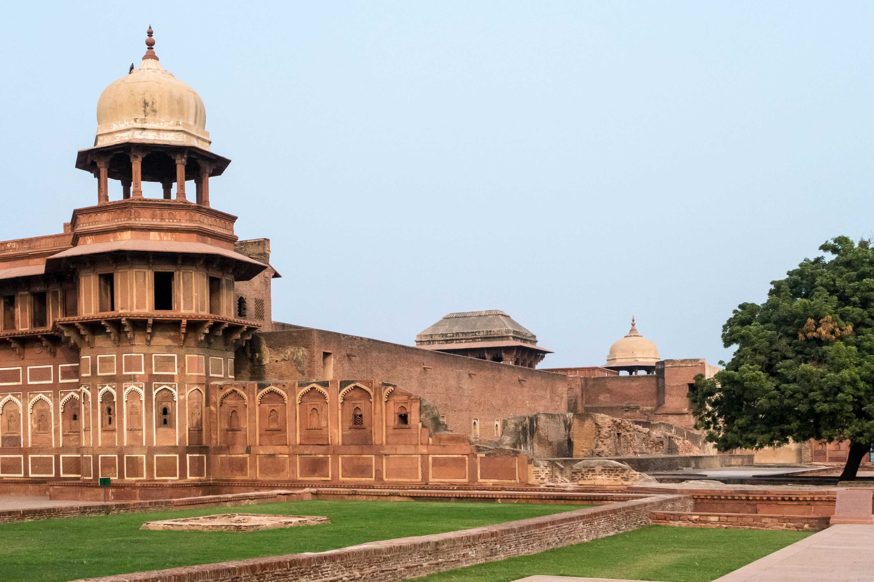 The image showcases Agra Fort in India. Visible are historic red sandstone structures, arched entrances, and an ornate dome. The fort's walls are interspersed with detailed carvings and windows. Lush green lawns and a large tree add contrast to the ancient architecture under India's clear sky.