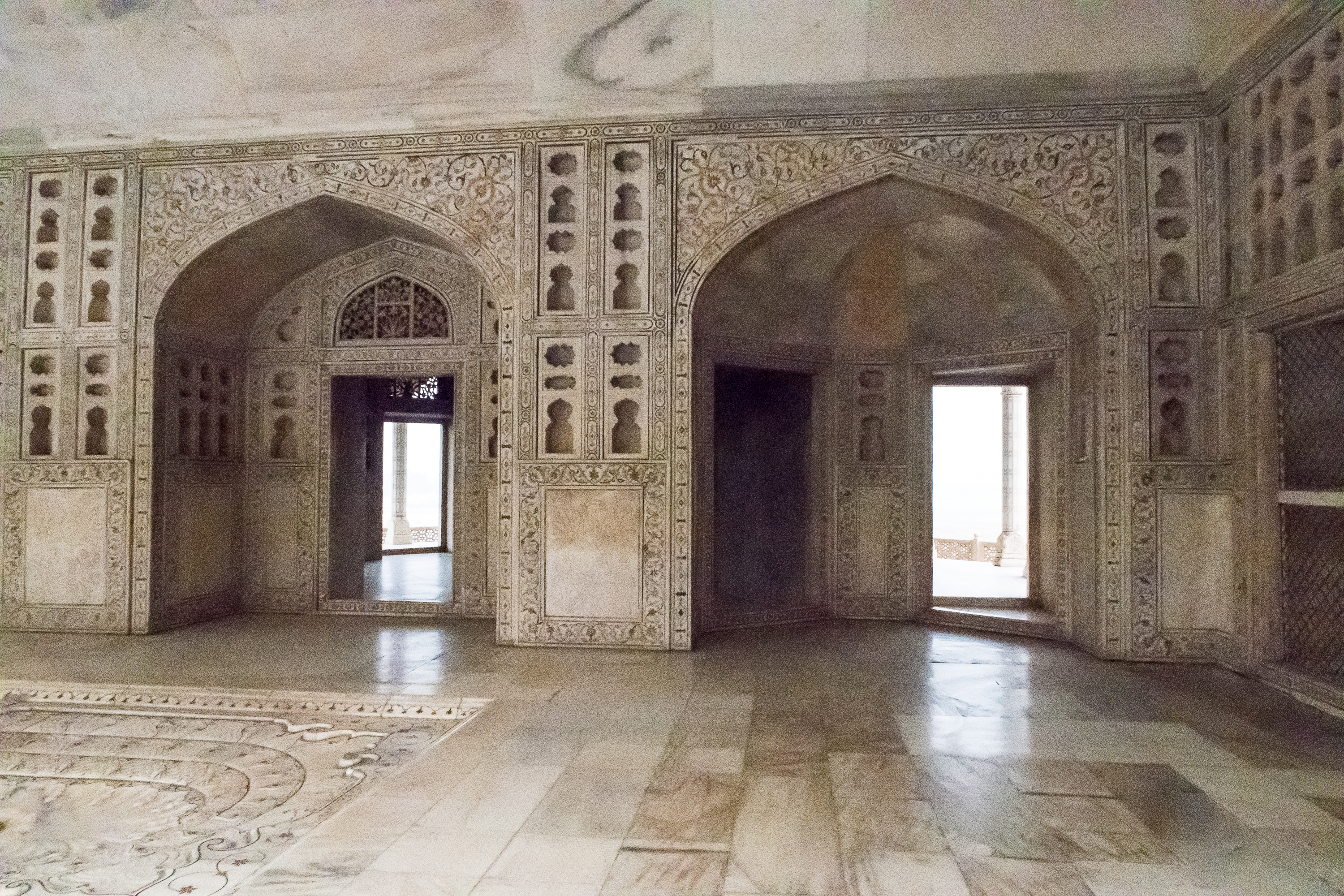 A historic stone room adorned with intricate carvings covers the walls and archways, reminiscent of ancient India. The floor exhibits a detailed, decorative pattern. Light filters in through the doorways and windows, illuminating the delicate craftsmanship of the architecture.