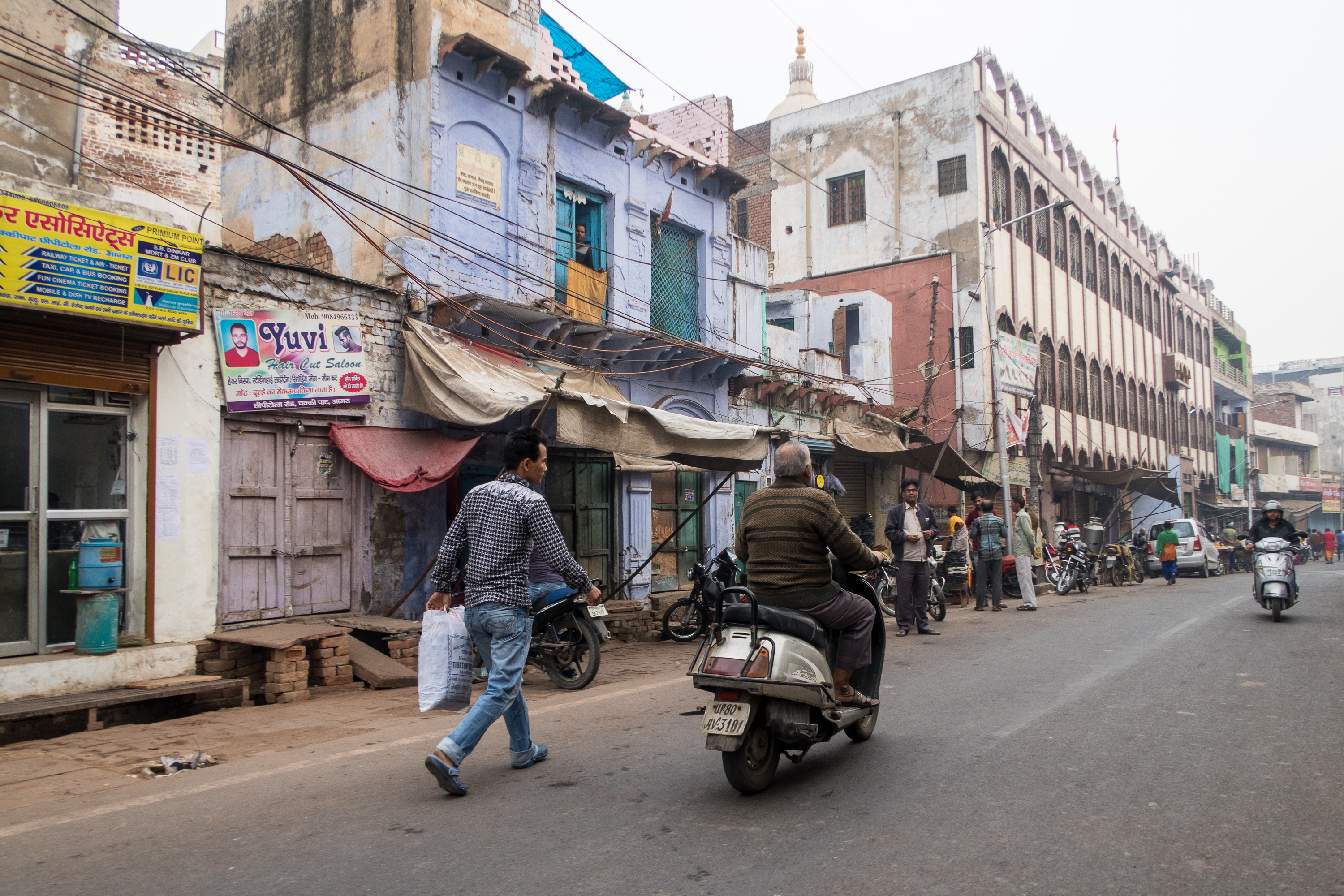 A busy street scene in India with people walking and riding bikes and scooters. The street, quintessentially Indian, is lined with various shops and old buildings, some with peeling paint and advertisements. Electrical wires crisscross overhead. Several vehicles are parked along the roadside.