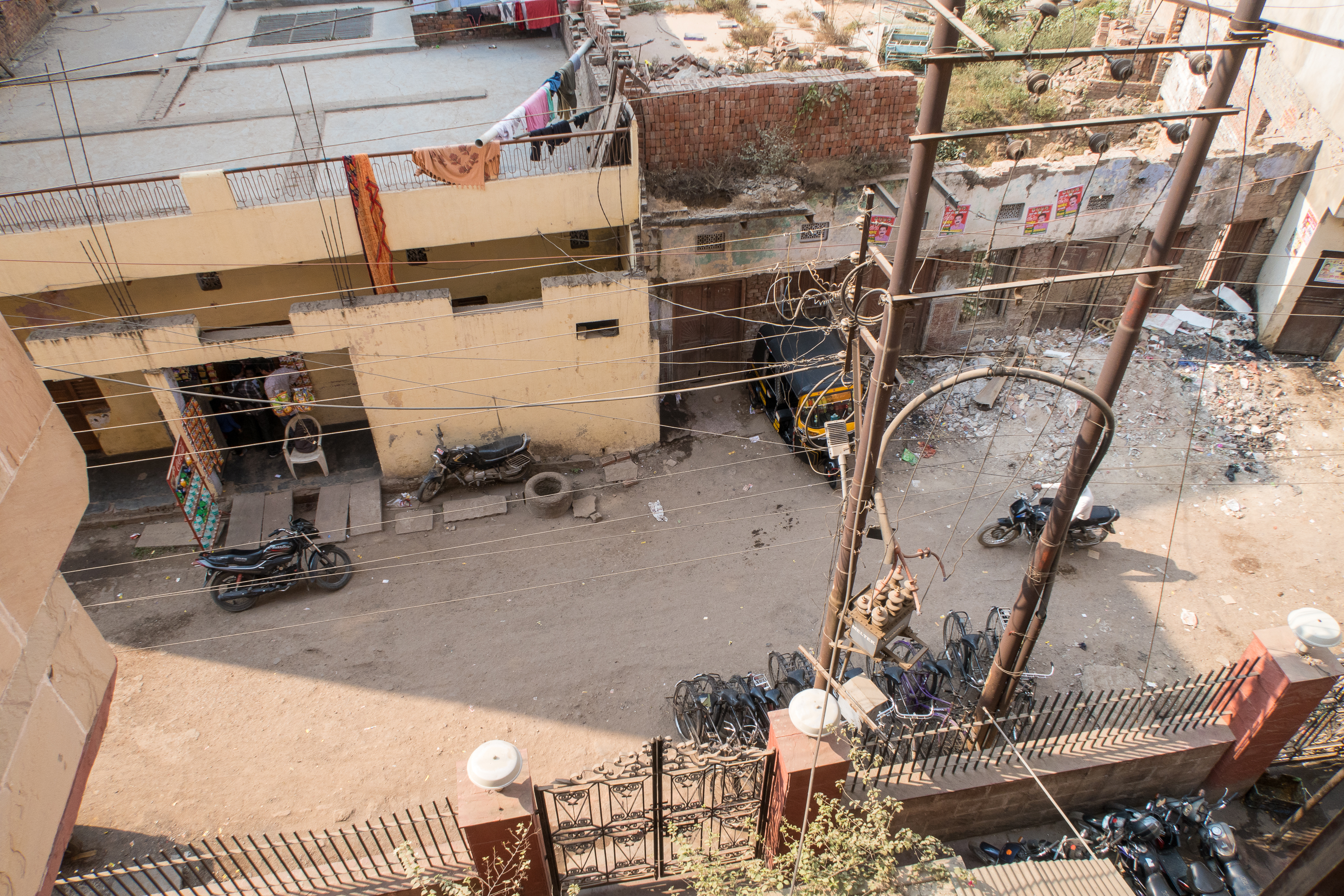 An aerial view of a dirt street in India, bordered by a yellow, multi-storied building with balconies holding clotheslines. The street is lined with parked motorbikes and an auto rickshaw. On the opposite side, a pile of garbage and rubble sits near a brick wall. Power lines crisscross above the scene.