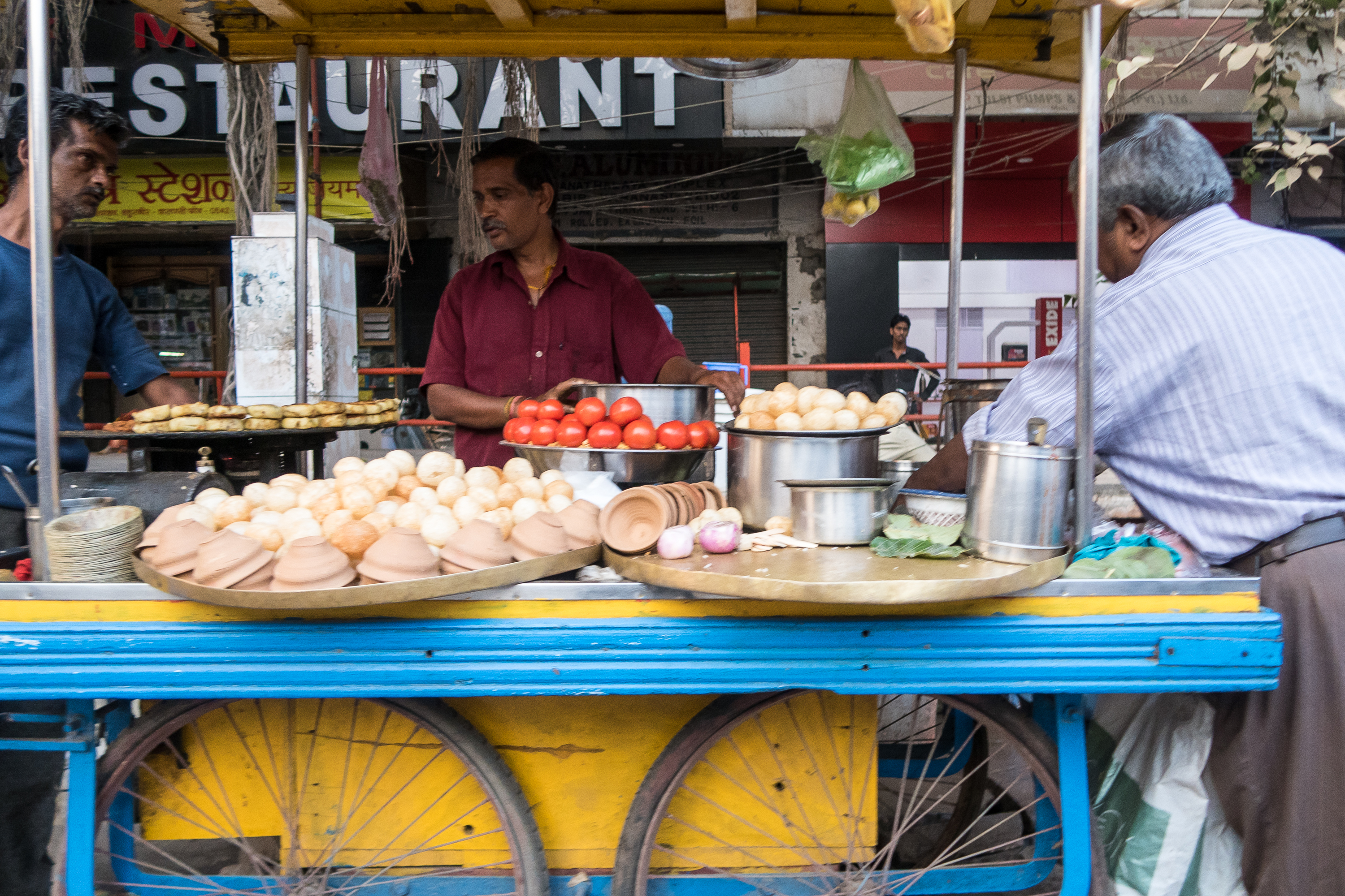 A street vendor in a maroon shirt stands behind his cart selling various food items, including tomatoes, bread rolls, and spices. The yellow and blue cart with large wheels adds vibrant charm to the bustling Indian street. Two customers are partially visible on either side, against a backdrop of restaurant signs and shuttered shops.