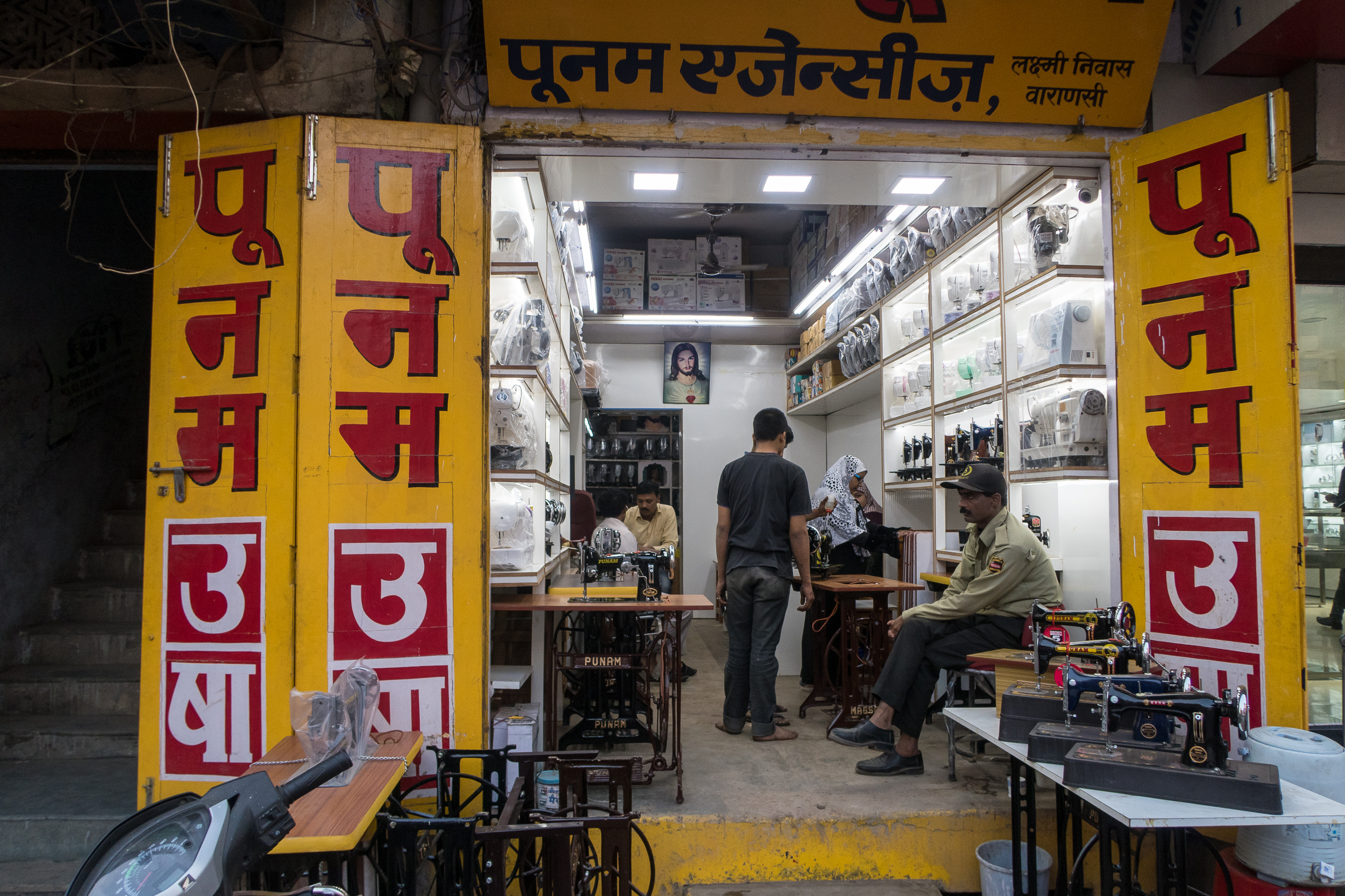 A sewing machine shop with yellow signage and red lettering. Inside, a person stands behind a counter displaying sewing machines, while another sits and one appears to be observing. Various models line the well-lit shelves. An individual, possibly a security guard, is seated outside the shop in India.
