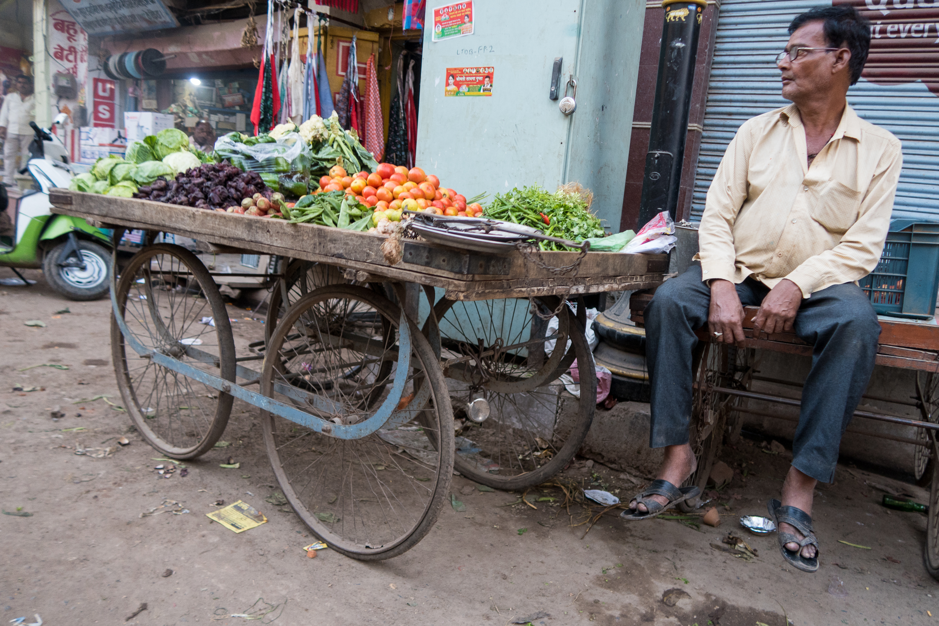 A street vendor sits on a bench beside his wooden cart filled with various fresh vegetables, including tomatoes, greens, and root vegetables. The scene is set in a bustling market area in India, with shops in the background. The ground is littered with debris, and a scooter is parked nearby.