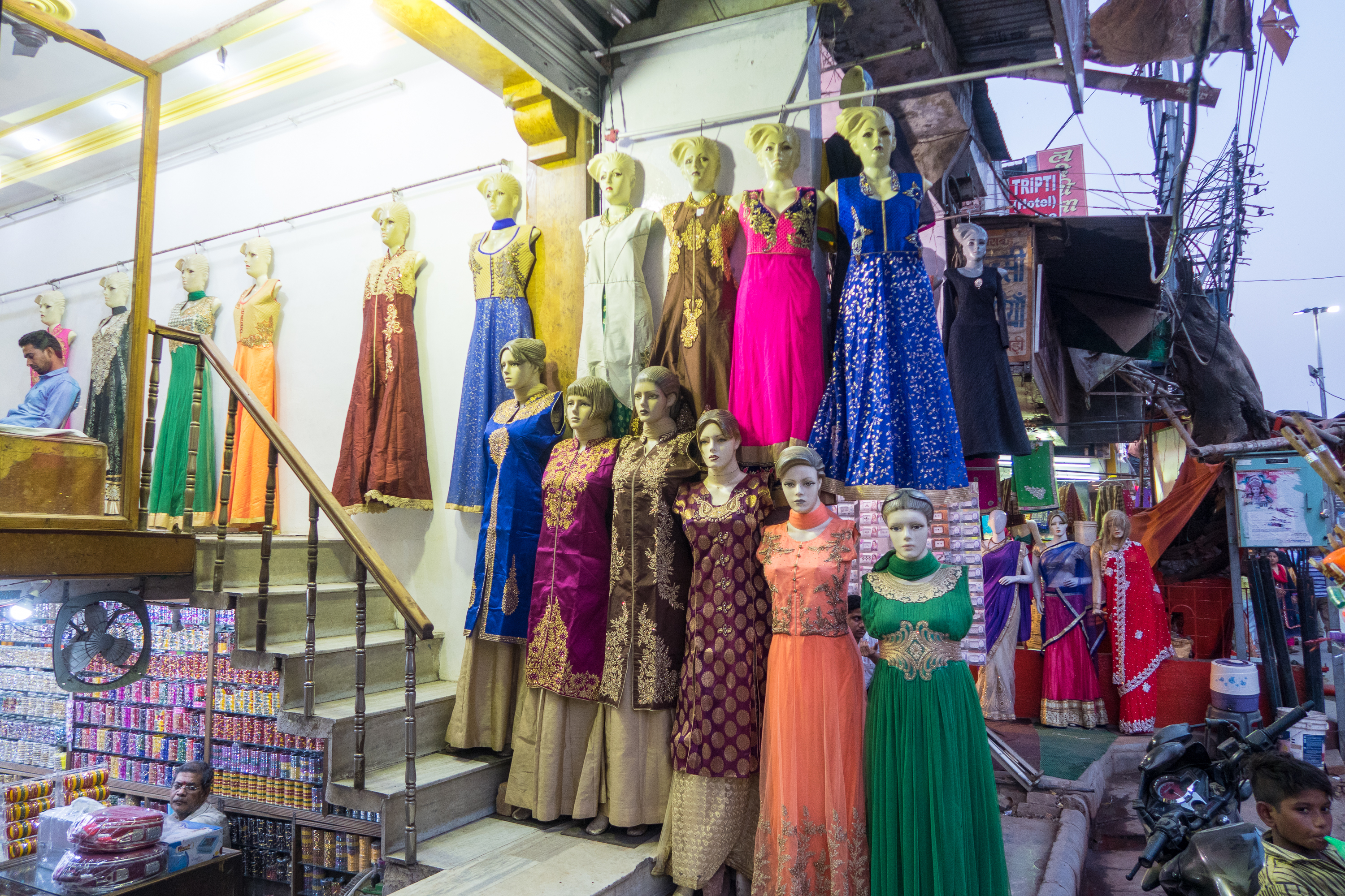 The image shows the front of a clothing shop in India, displaying an assortment of colorful traditional dresses on mannequins. Located in a busy market area, the mannequins are arranged on tiered steps, showcasing various styles and vibrant colors, with potential customers and other market stalls visible in the background.