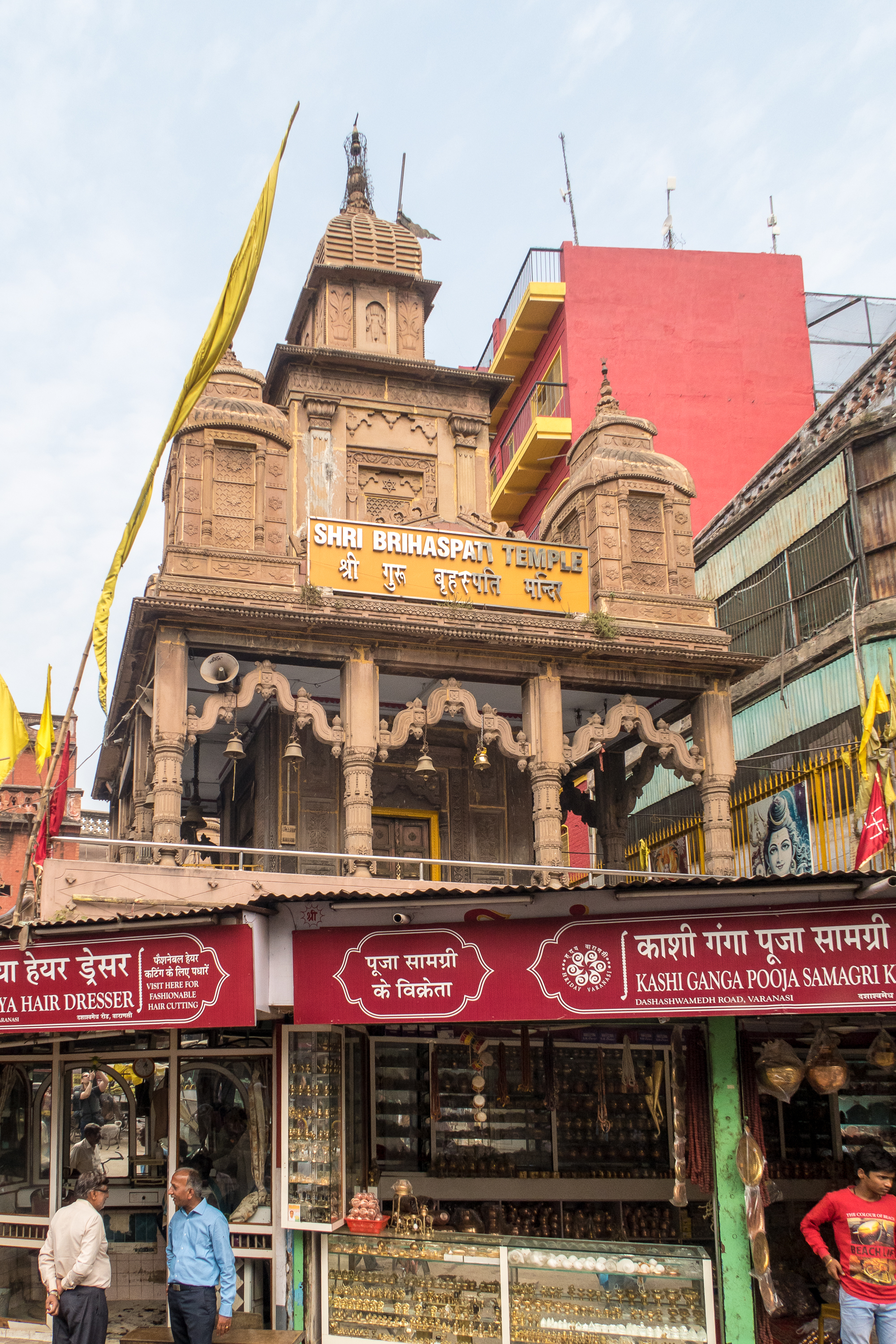 A bustling street in India with a historic temple, the Shri Brihaspati Temple, prominently displayed above shops, including a hairdresser and a puja samagri store. The temple features intricate carvings and multiple spires. Flags and signs in Hindi adorn the buildings, with people walking nearby.