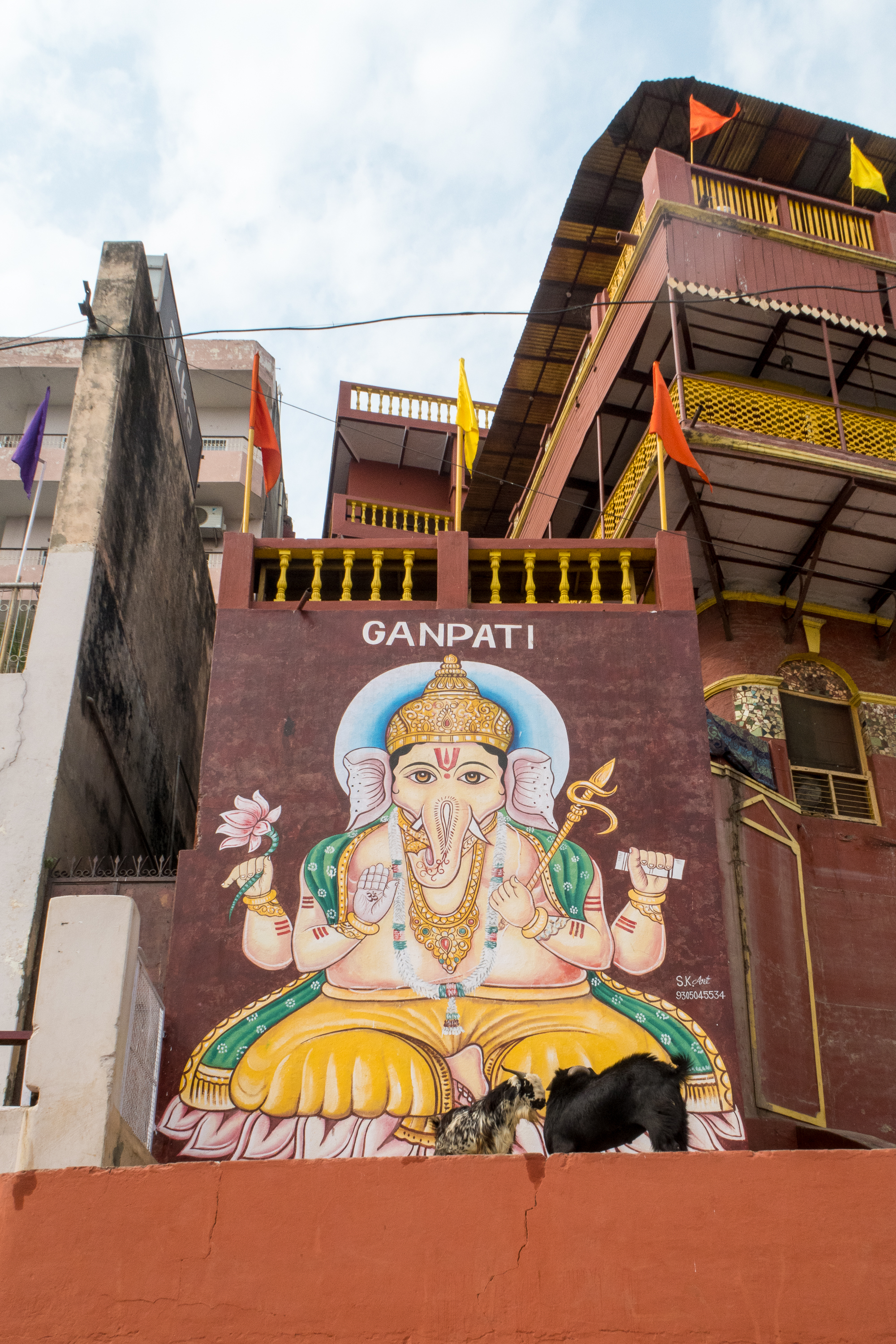 A large mural of the Hindu deity Ganesha is painted on a building wall in India. The artwork features Ganesha seated, with multiple arms holding various objects. Above the mural, the word "GANPATI" is written. The building is decorated with colorful flags and sits under a partly cloudy sky.