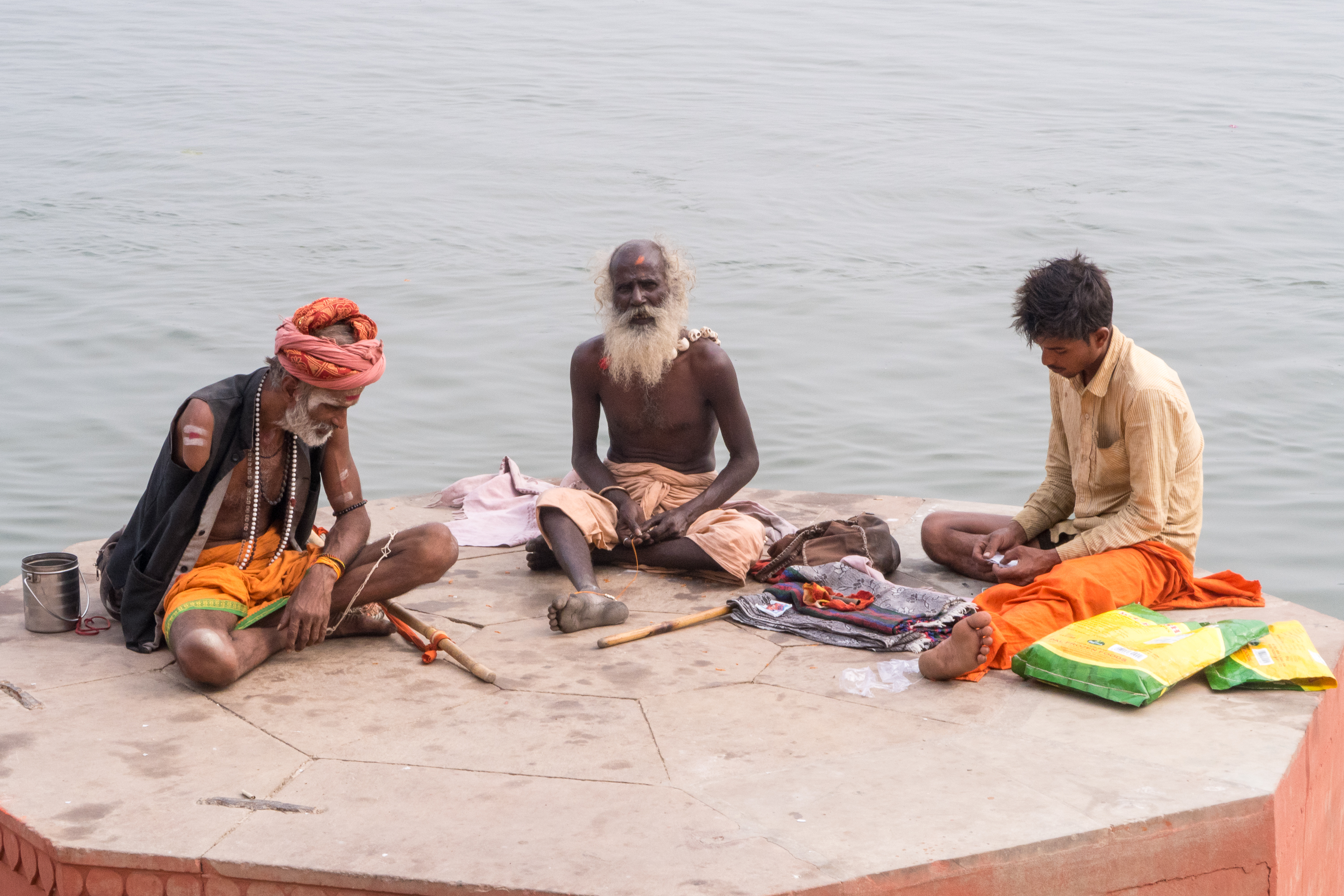 Three men sit cross-legged on a hexagonal concrete platform by a body of water in India. The man on the left has an orange headwrap and wears beaded necklaces. The central man, older with a white beard, sits with a piece of cloth on his lap. The third man, in beige and orange, is surrounded by various items.