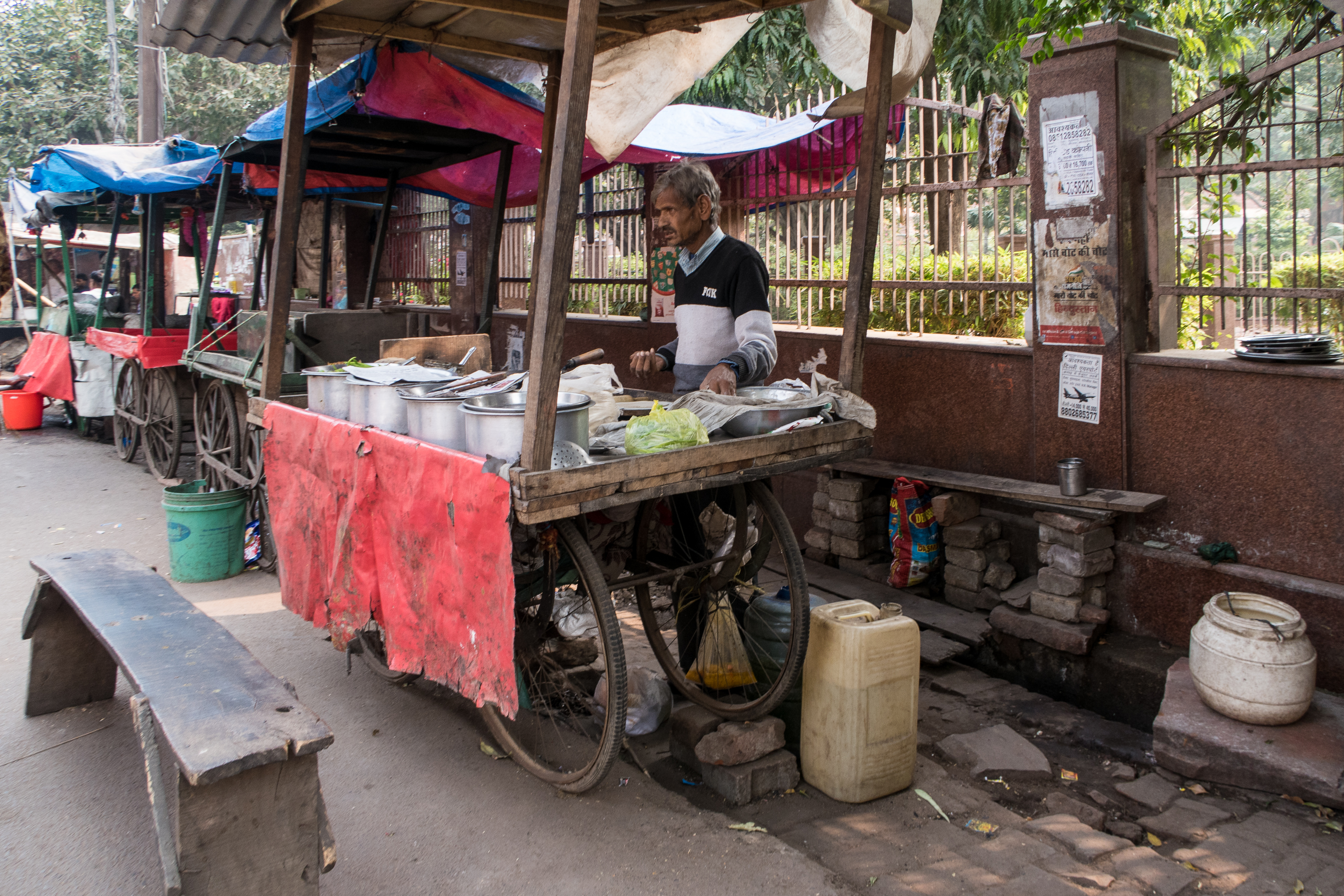 A street vendor in India stands behind a makeshift cart draped with a red cloth, surrounded by various cooking pots and containers. The stall is set up along a fence adorned with signs. A bench is placed in front, and on the ground lie a plastic canister and a white container.
