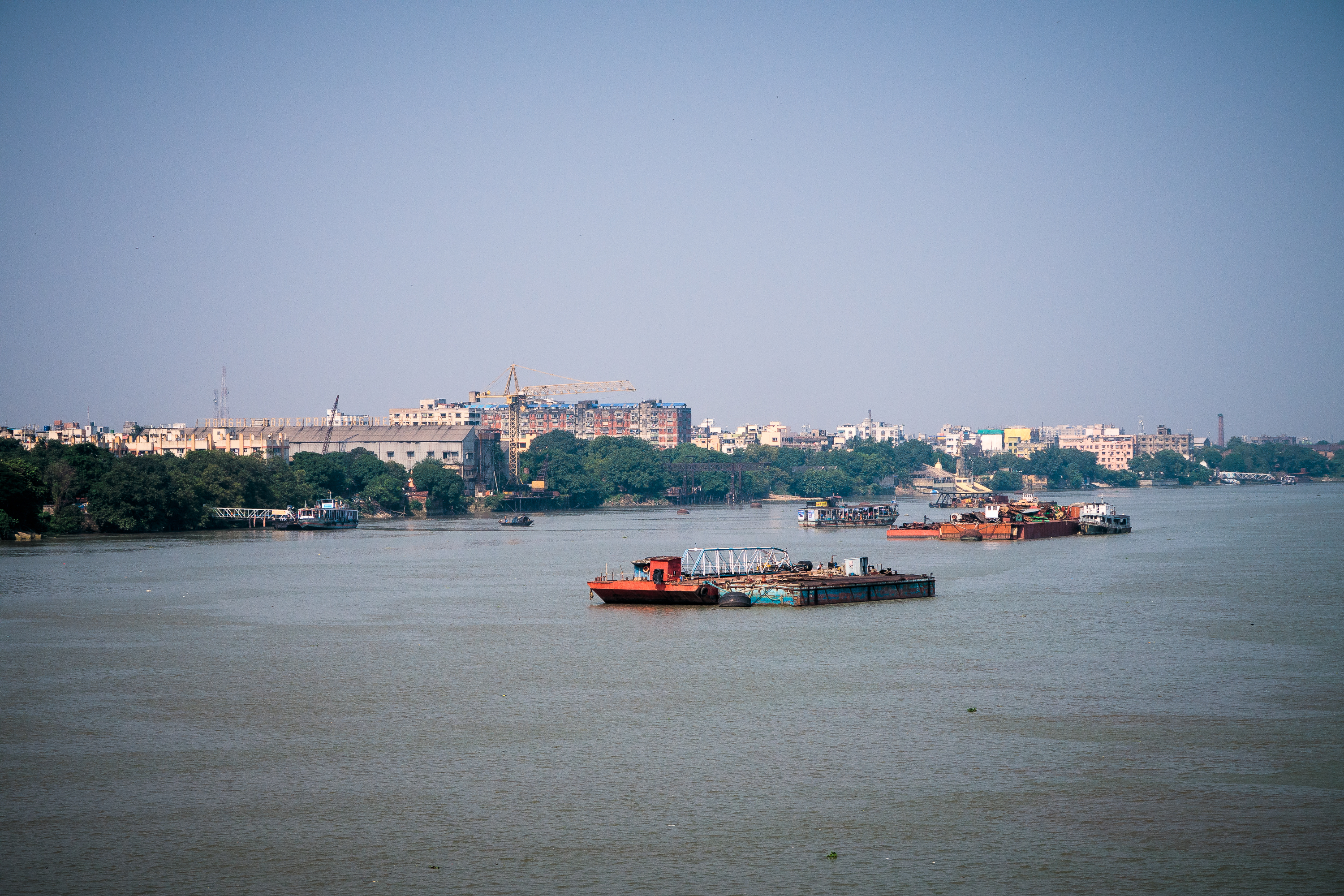 A river with several boats and barges floating on its calm waters under a clear blue sky in India. A distant shoreline features a mix of green trees and urban buildings, including some under construction. The backdrop includes various architectural structures blending into the city's landscape.