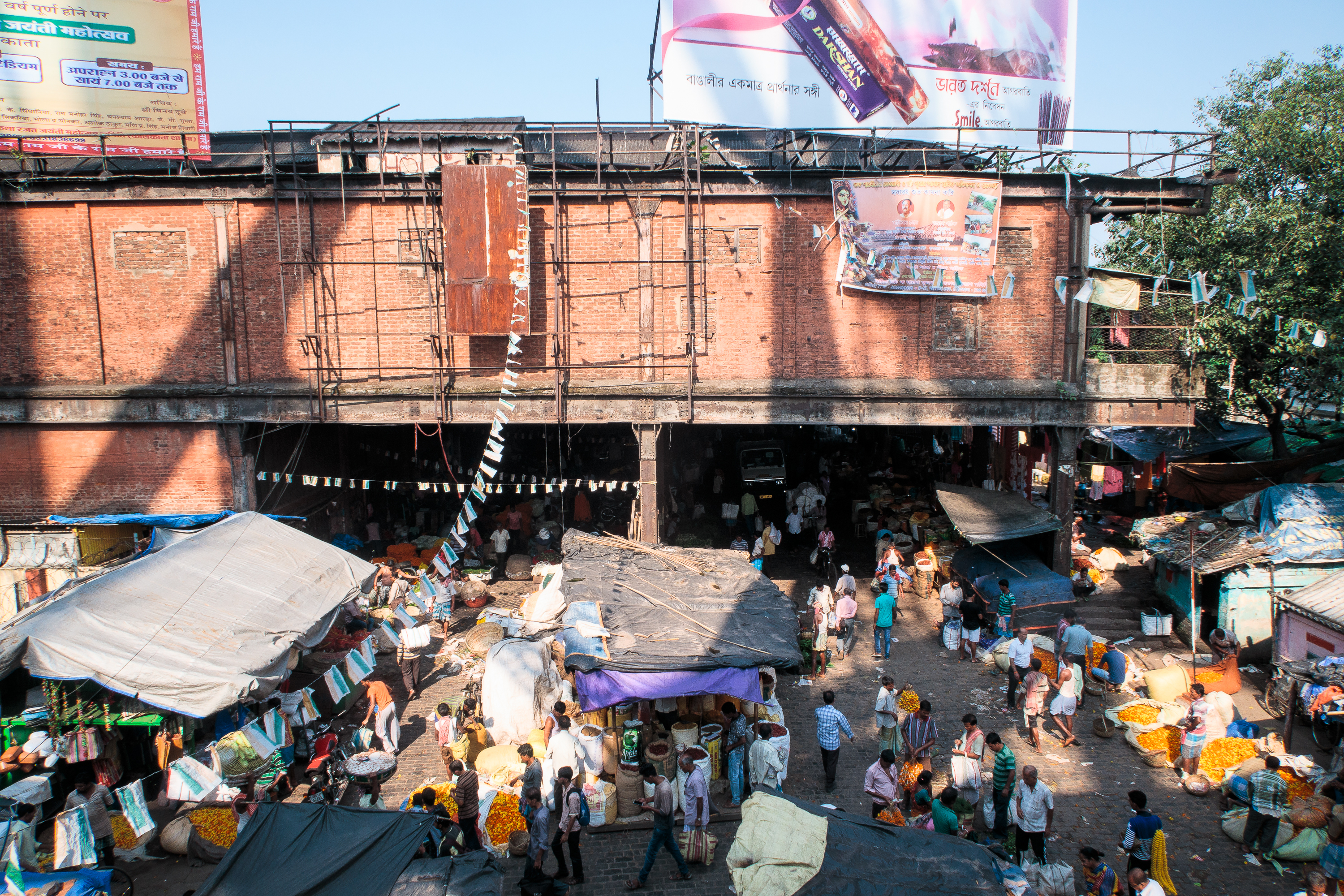 A bustling outdoor market scene in India with numerous vendors and customers. Stalls covered with tarps are laden with produce, especially yellow and orange flowers. The market is set against a backdrop of a large, old brick building with multiple billboards and some hanging signage, under a clear sky.