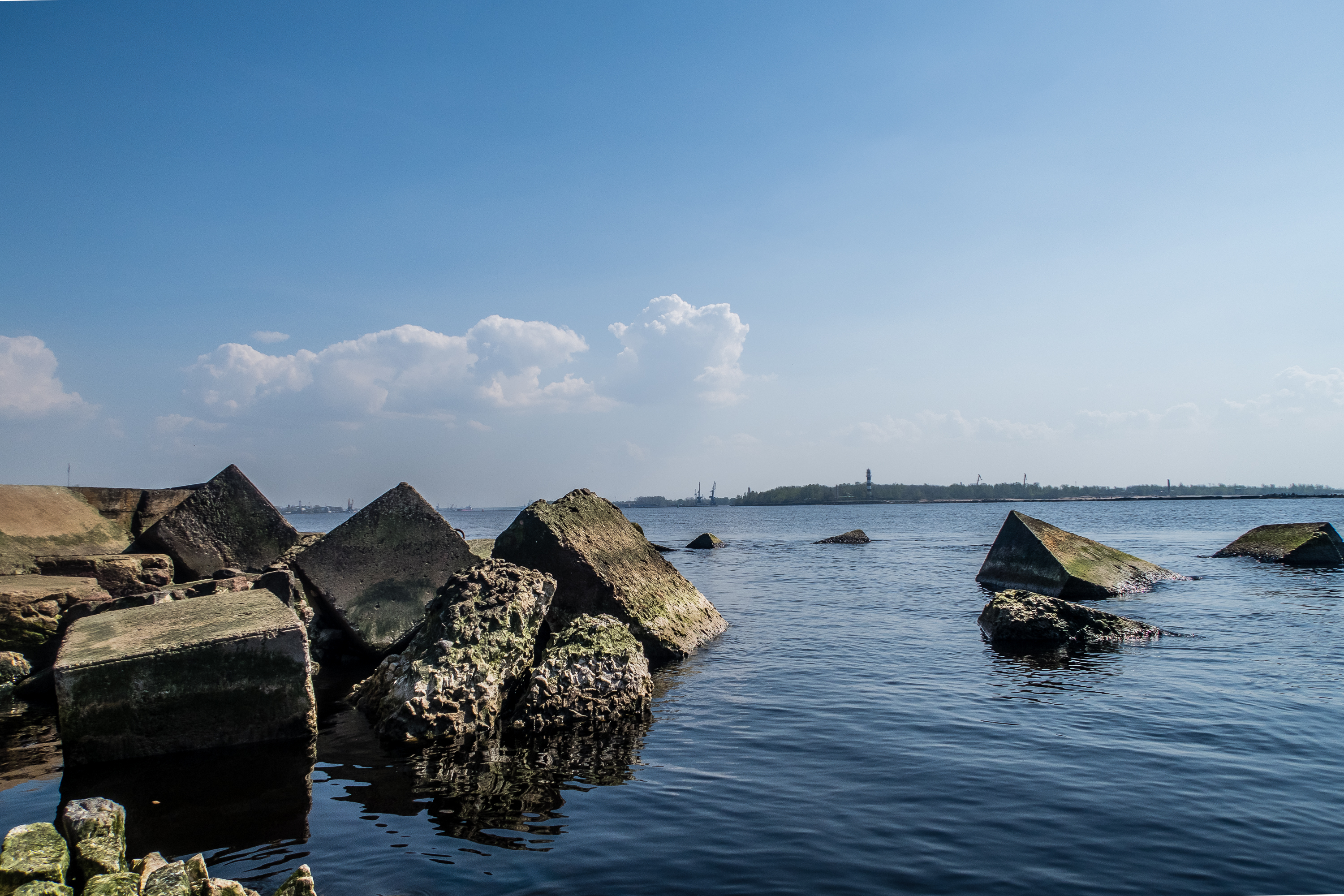 Large, moss-covered concrete blocks sit partially submerged beside a jetty in a calm body of water under a clear blue sky with scattered clouds. In the distance, a strip of land with some trees and structures is visible on the horizon. The scene is peaceful and bright.