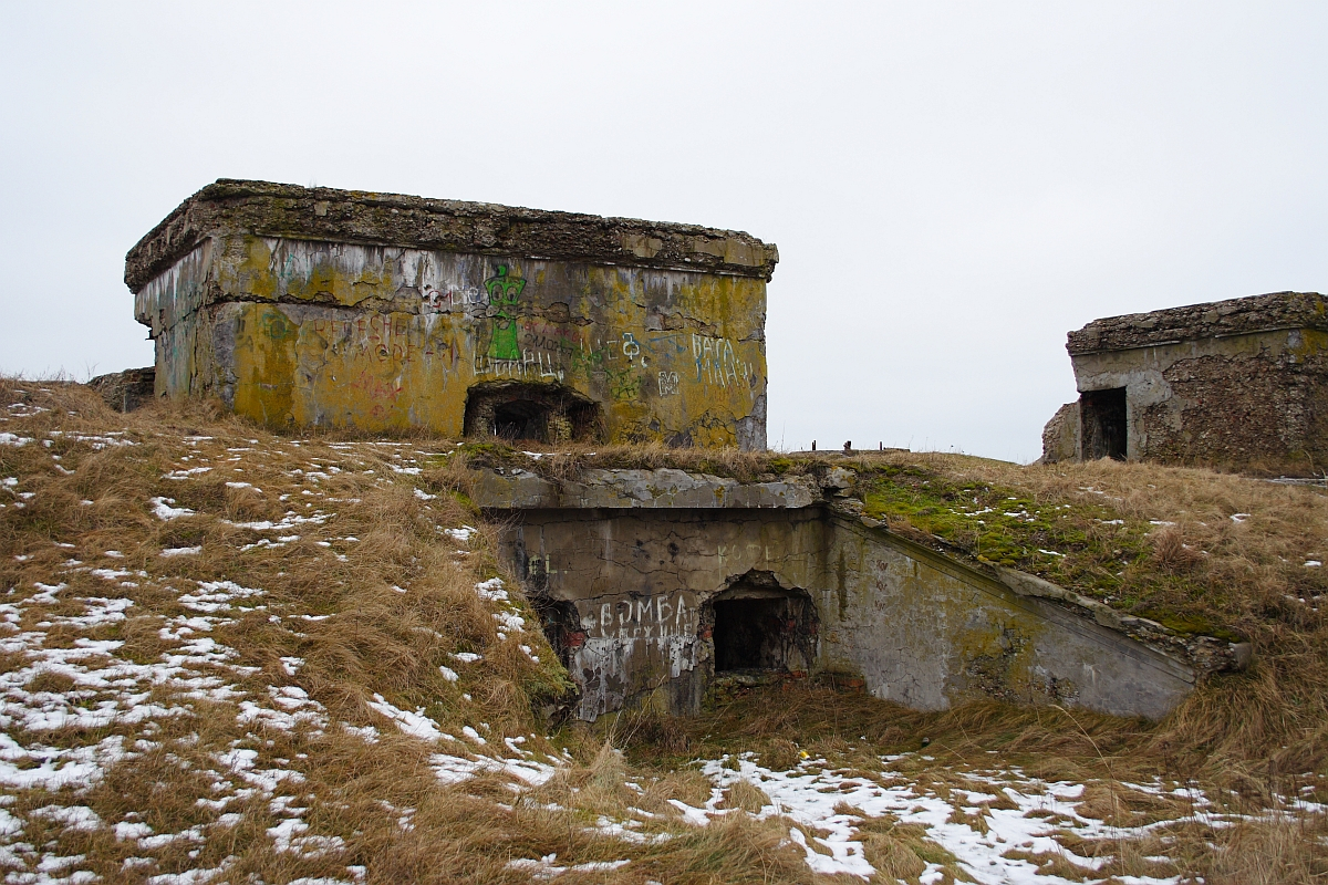 An abandoned concrete bunker complex stands amid a barren landscape with sparse snow patches in what was once the former military town of Karosta. The bunkers are weathered, covered in graffiti, and surrounded by dry grass. The overcast sky enhances the desolate atmosphere, highlighting the deteriorating structures with multiple entry points.