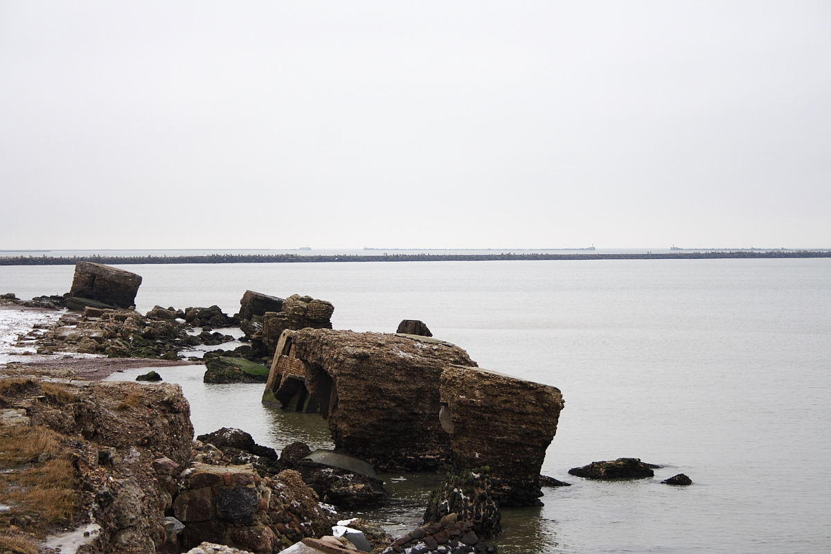 A serene coastal landscape featuring scattered rocky remnants along the Karosta shore, with calm waters extending to a distant horizon under an overcast sky.