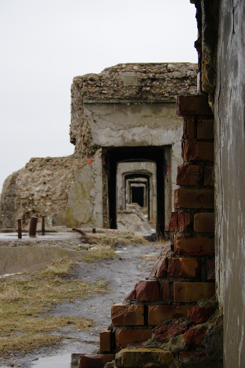 A view through multiple archways of the weathered, abandoned Karosta building. The walls are crumbling, revealing exposed bricks and concrete. The ground is uneven, with patches of grass and rubble. The sky is overcast, casting a dull light over the decaying, historic structure.