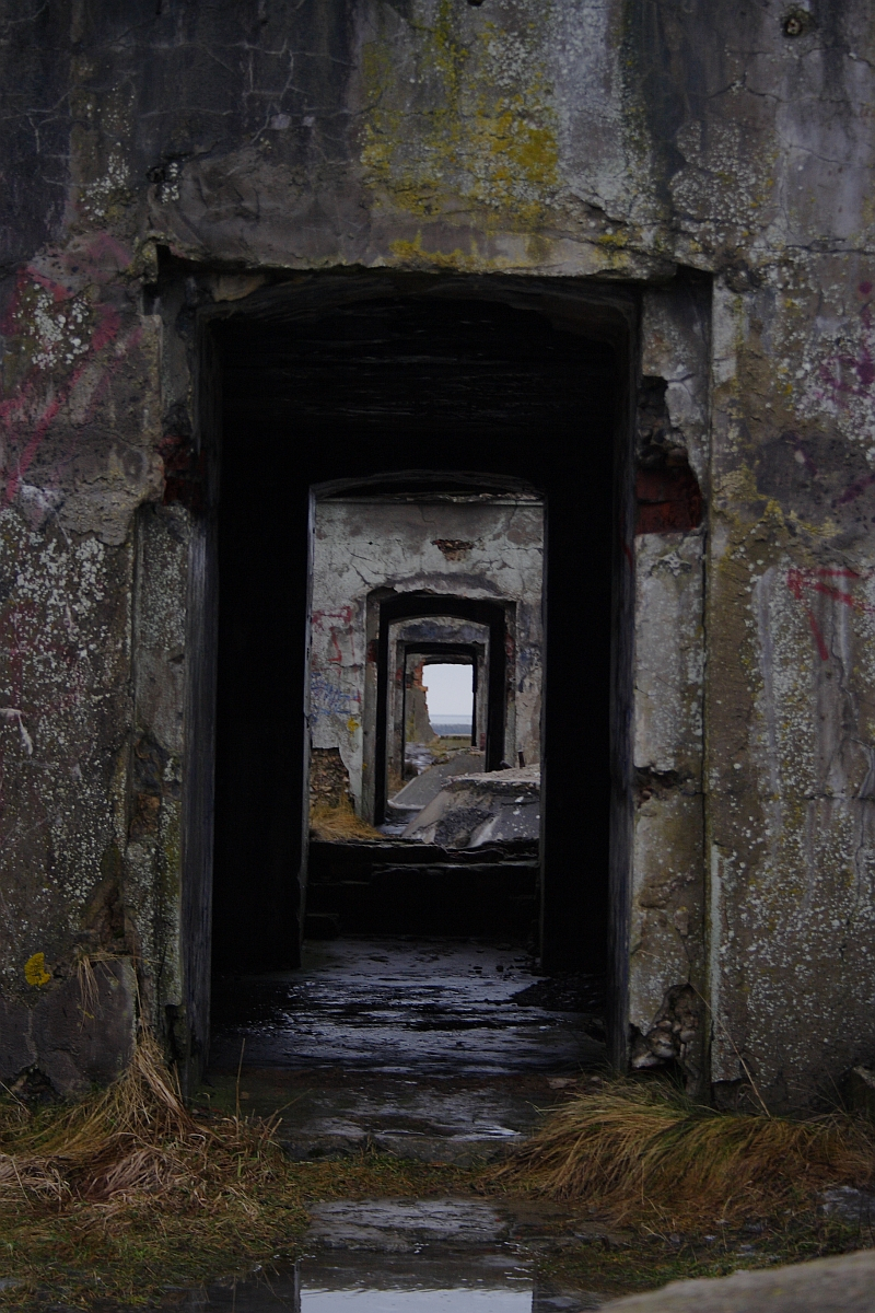 A series of dilapidated doorways creates a haunting tunnel effect in the old, abandoned Karosta building, with layers of graffiti adorning the decaying walls, and nature slowly reclaiming the once