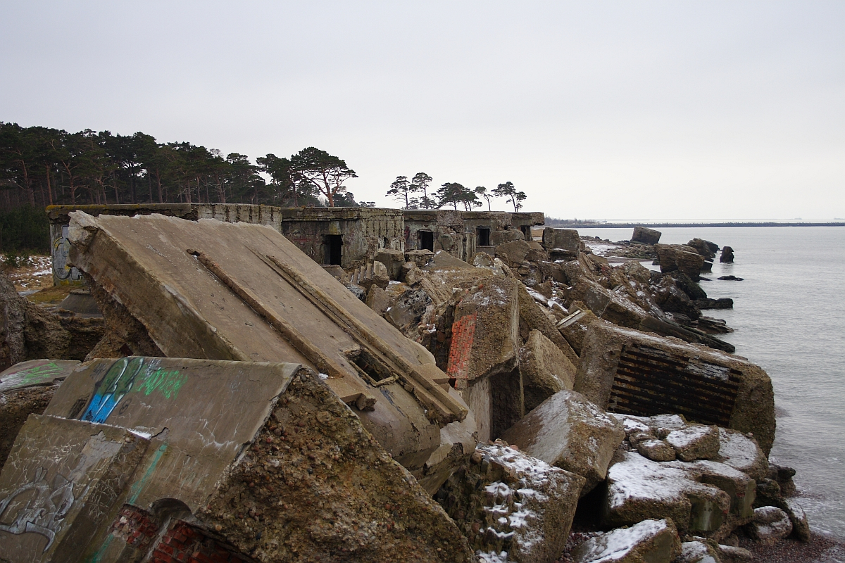 Ruins of an old coastal fortification in Karosta, with dilapidated concrete structures scattered along a snowy beach, trees in the background, under an overcast sky.