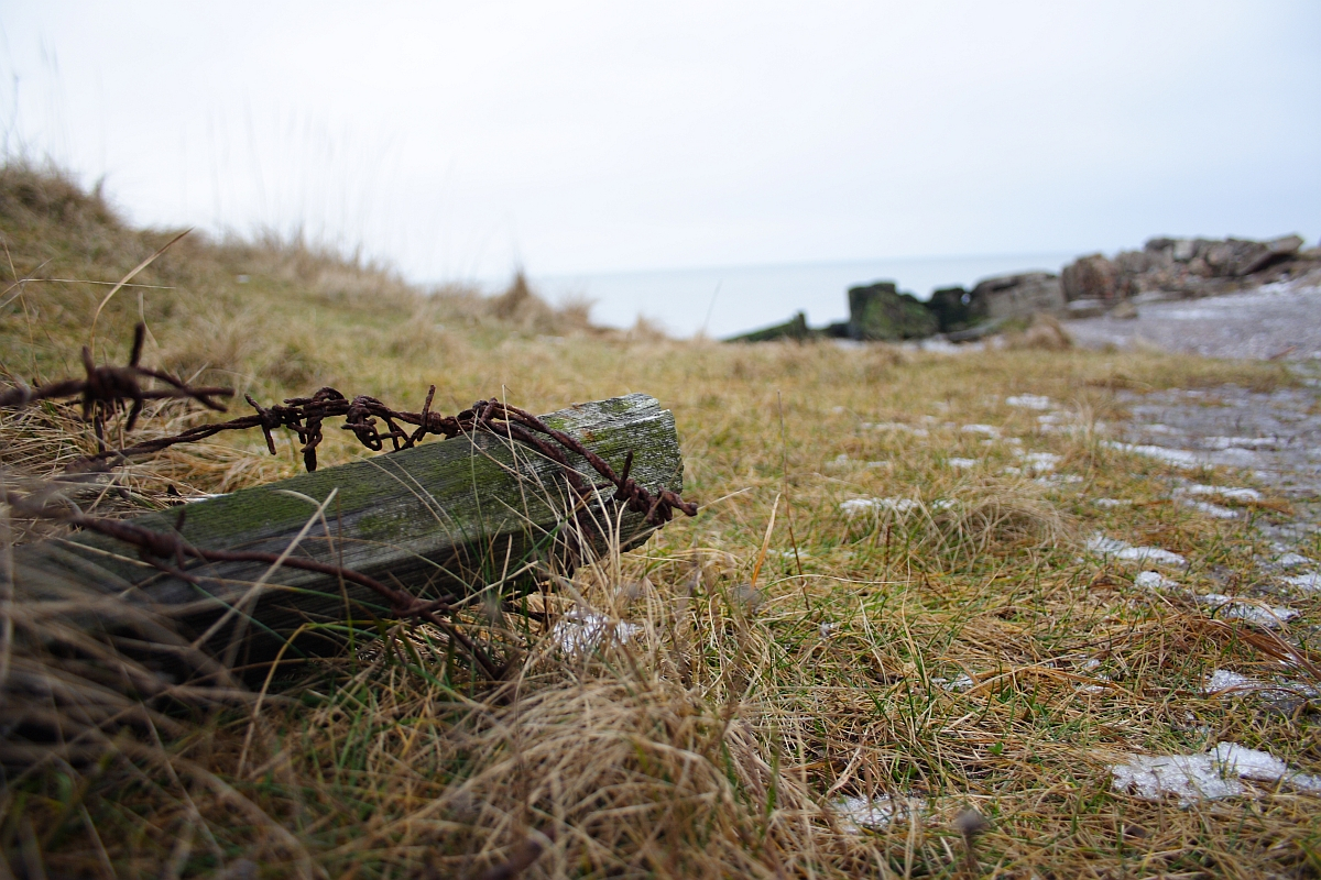 A weathered wooden post wrapped in rusty barbed wire lies in the foreground, surrounded by dry grass and sparse patches of snow. In the background, more wooden posts and rocks scatter across the desolate Karosta coastline, with an overcast sky and calm sea visible on the horizon.