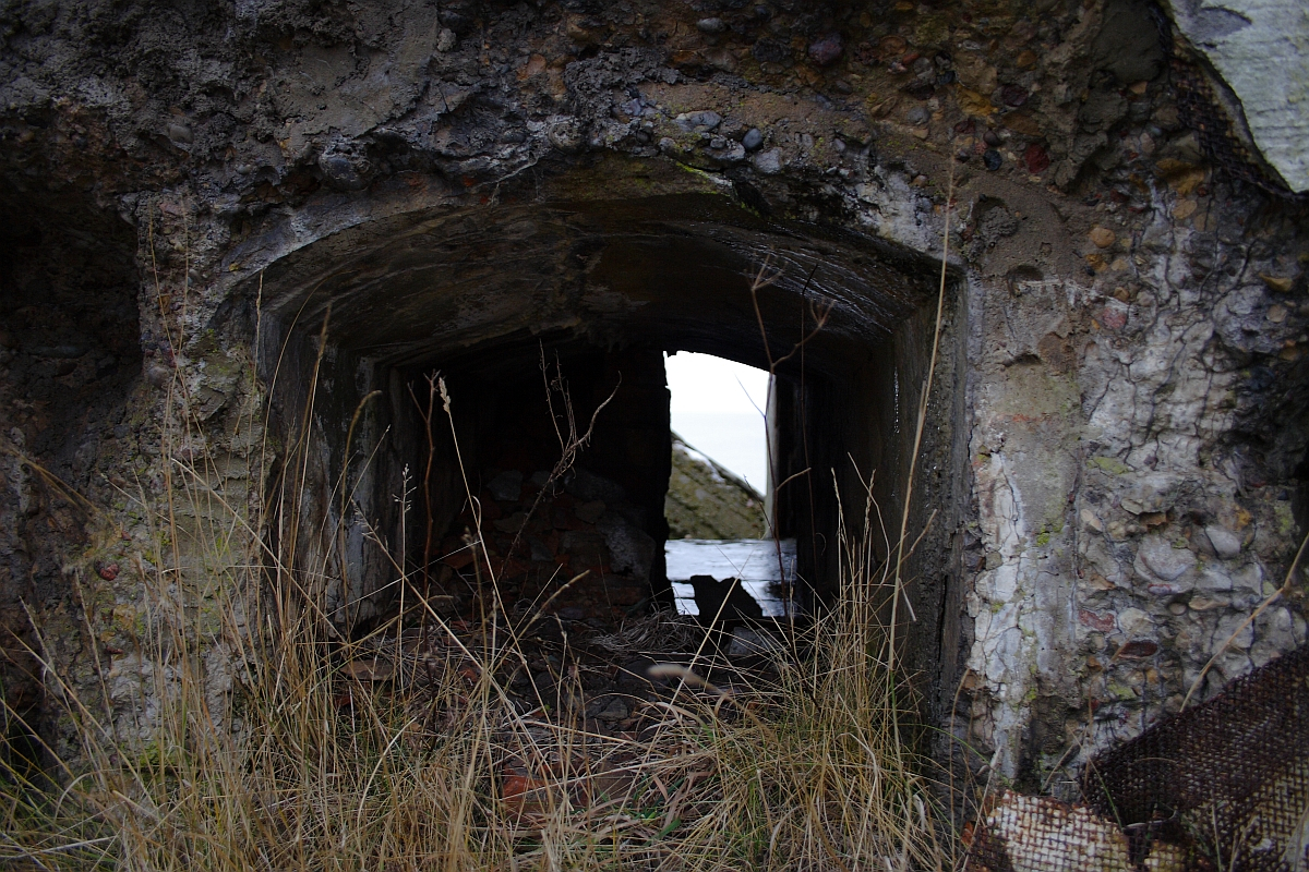 An abandoned bunker entrance in Karosta, overgrown with wild grass, the dark interior hinting at a forgotten past, set against a bleak sky.