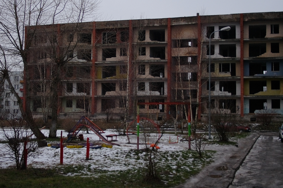 A bleak winter scene in Karosta showing an abandoned, dilapidated building with broken windows behind a desolate playground with scattered snow patches, evoking a sense of neglect and desolation.