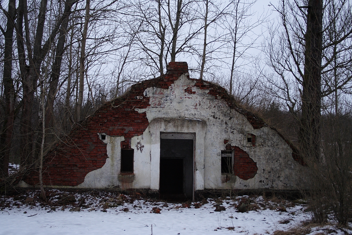 An abandoned, dilapidated structure in Karosta with a crumbling red brick facade and a derelict white plaster front stands forlornly amidst a leafless winter forest, with remnants of snow