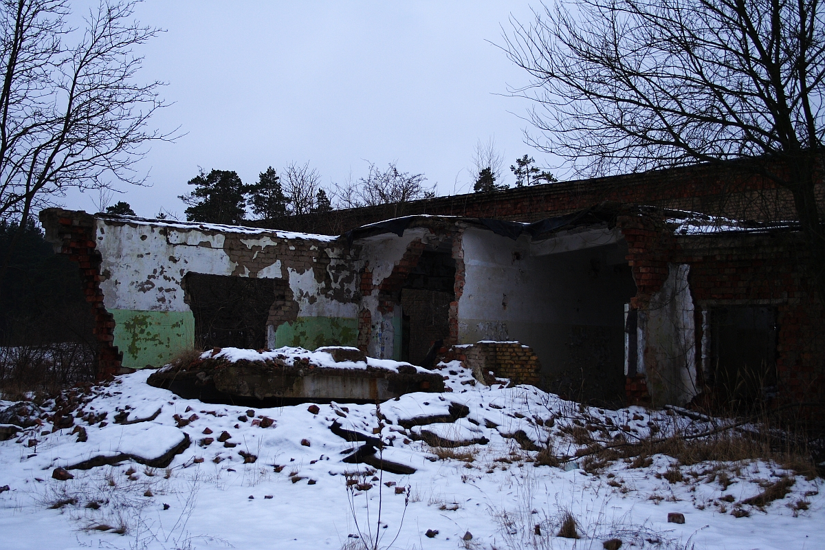A dilapidated brick building in Karosta stands in wintery decay, its walls crumbling under the weight of time, partially clad in remnants of green and white paint, the snow gently blanketing
