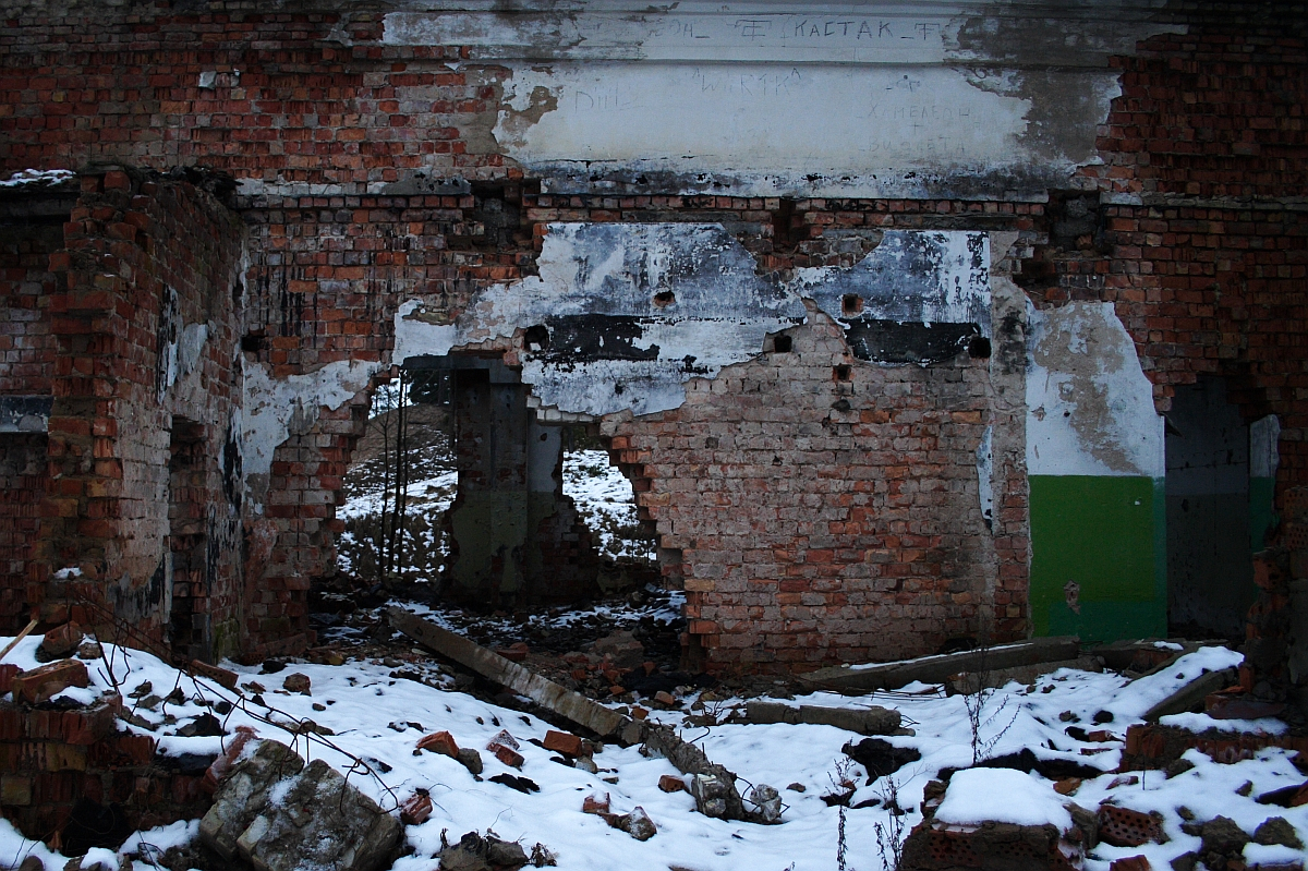 A dilapidated brick structure in Karosta with remnants of white plaster, featuring a graffiti-tagged wall and a snow-dusted ground, stands as a testament to decay and abandonment.