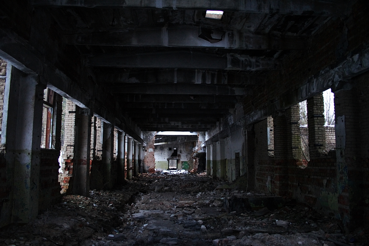 An eerie, dilapidated interior of Karosta's abandoned building, showcasing crumbling brick walls, scattered debris, and a desolate hallway leading into darkness, under the remnants of a once-sturdy concrete