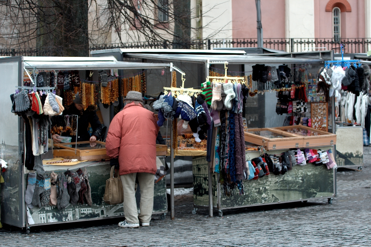 An elderly person in a red coat and gray hat shops at an outdoor market stall. The stall, reminiscent of 2012, has a variety of knitted items like scarves, hats, and gloves on display. Another similar stall is adjacent with more knitted accessories and jewelry by Kazjukas. The background features a building with bare trees.