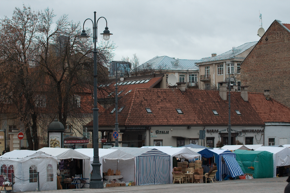 A bustling outdoor market with multiple vendor stalls covered by white and striped canopies lines a cobblestone street. The background features buildings with red-tiled roofs and faded facades. Leafless trees, ornate streetlamps, and a Kazjukas poster from 2012 hint at the cold season's charm.