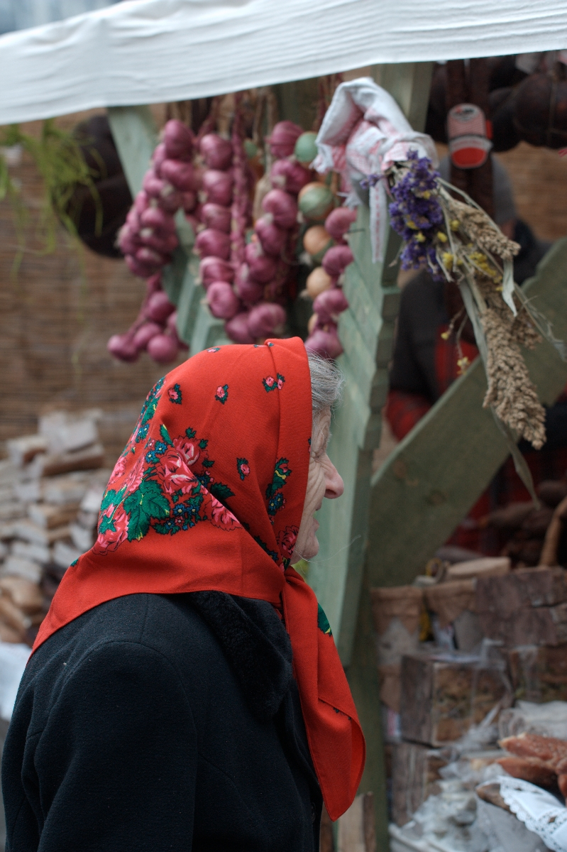 An elderly woman wearing a black coat and vibrant red floral headscarf stands in front of a wooden stall at the 2012 Kazjukas festival. The stall is adorned with hanging bunches of dried red onions and assorted herbs, while the background features stacked firewood and partially visible textiles.