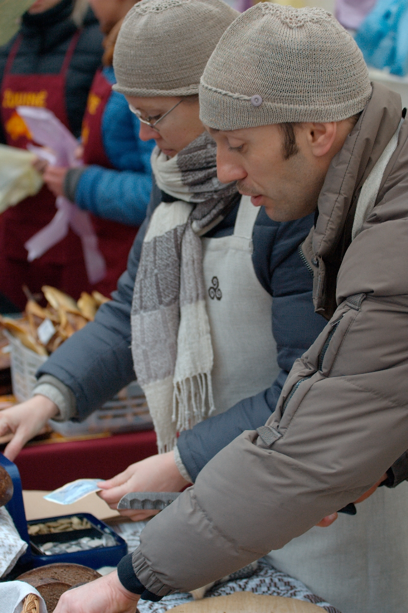 Two people, dressed warmly in coats, knitted hats, and scarves, serve food at an outdoor market. They are focused on their tasks, handling trays and plates. A third person in the background, also warmly dressed, appears to be assisting. The scene is lively and bustling like Kazjukas 2012.
