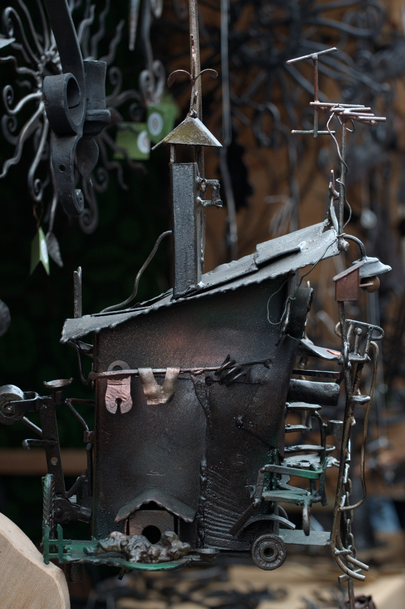 A metal sculpture by Kazjukas from 2012 depicts a whimsical, abstract house with a slanted roof, chimney, and attached wire details. The house blends geometric and organic forms, with metal elements resembling clothes on a line and twisted wirework suggesting branches or antennae. Background is blurred.