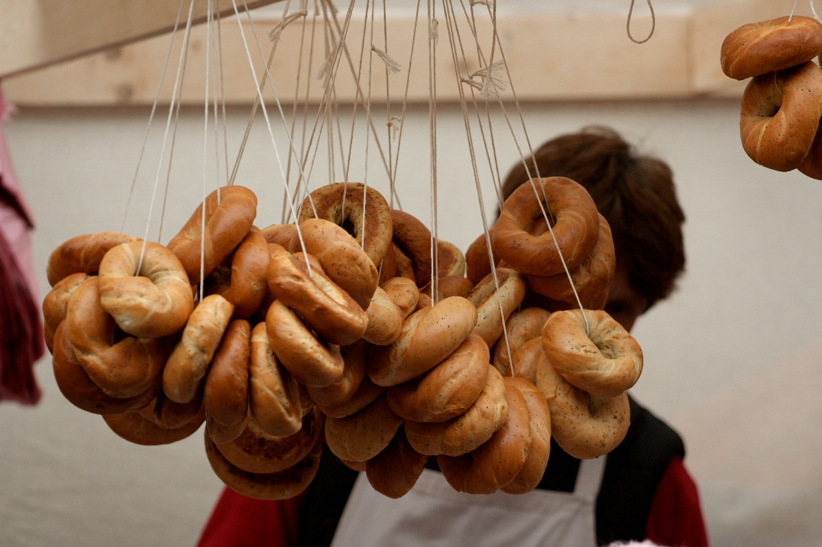 A variety of bagels hang by strings from the ceiling of a market stall. The bagels come in different shades of brown, indicating various types or levels of baking. A person with brown hair is partially visible in the background, focusing on the display, reminiscent of a scene from Kazjukas 2012.