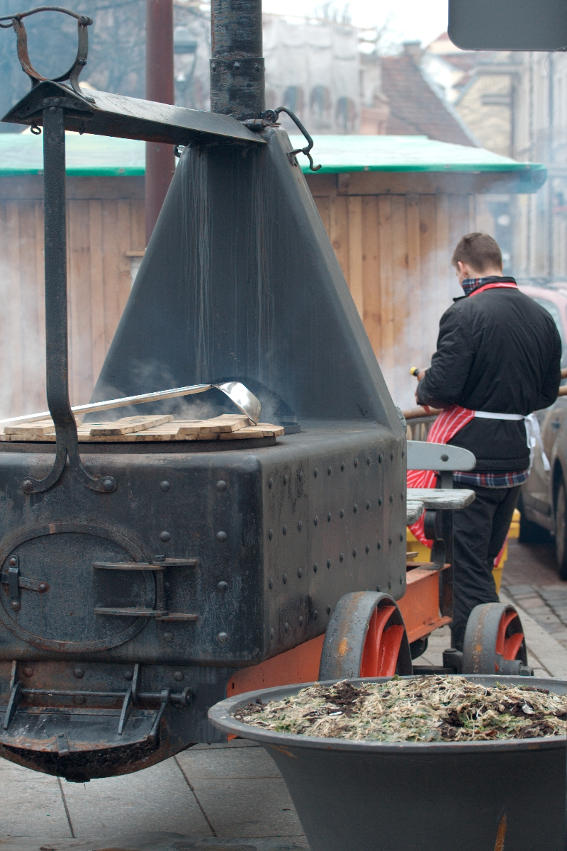 A person stands beside a large, mobile metal oven on wheels, likely used for cooking or smoking food outdoors. The individual, possibly Kazjukas, is wearing a black jacket and red apron, facing away from the camera. In the foreground, there's a bowl filled with what appears to be dry vegetation or herbs.