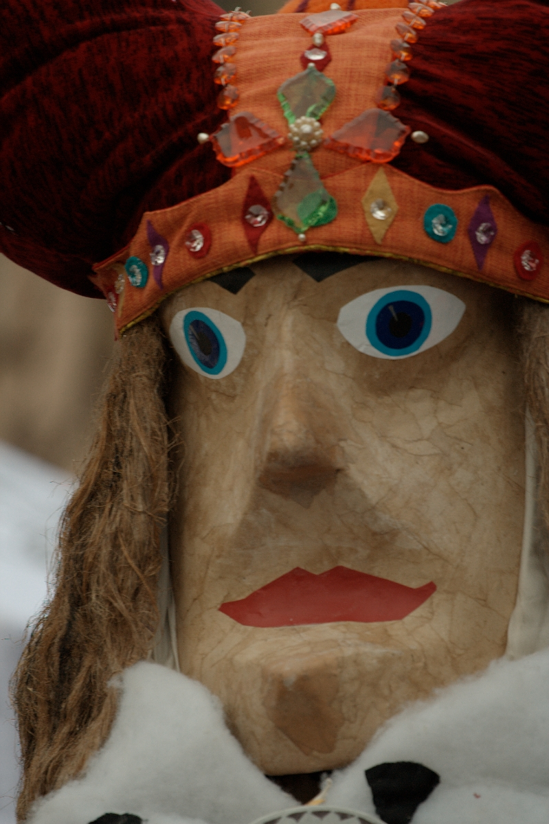 Close-up of a large wooden puppet with exaggerated facial features including bright blue eyes and red lips. The Kazjukas 2012 puppet has long hair and wears a headdress adorned with multicolored jewels and a large red, bejeweled top section. The headdress includes a decorative green diamond-shaped element.