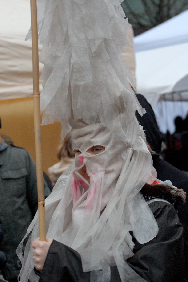 A person is dressed in a costume with layers of white, crinkly material covering their head and face, extending upwards to form a tall, pointed hat. The material has a slightly translucent appearance. They are holding a wooden pole and wearing a dark coat, reminiscent of the Kazjukas celebration in 2012, with other indistinct figures in the background.