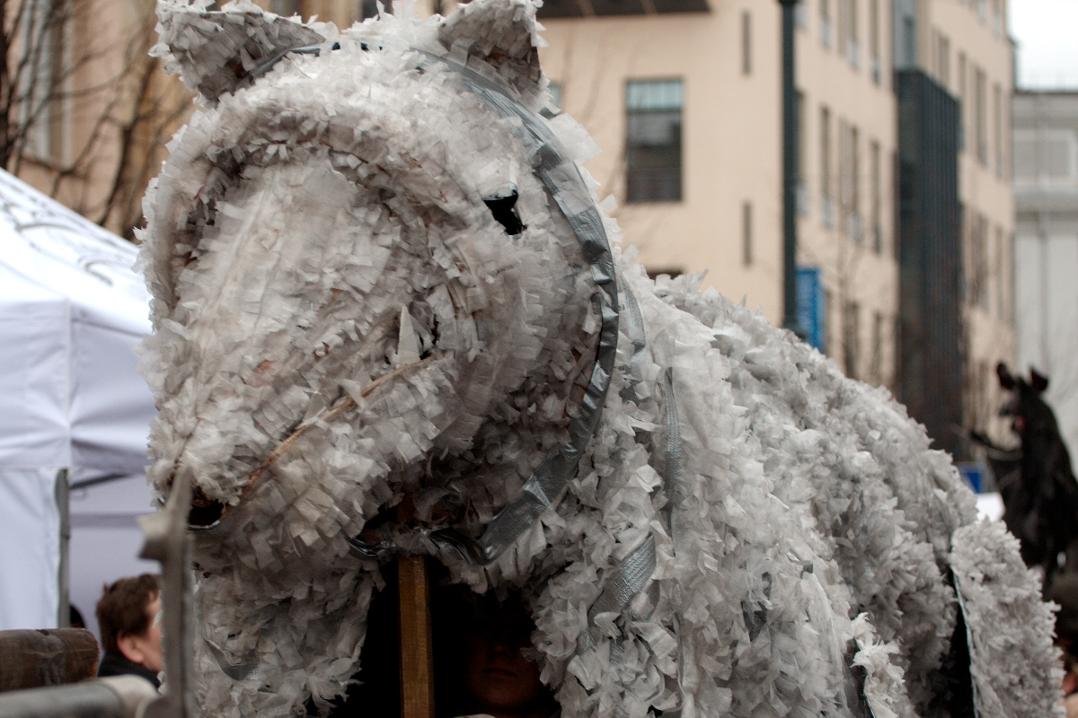 A large, gray, horse-shaped puppet covered in frayed fabric and supported by wooden poles is part of a lively street performance or parade. People can be seen underneath and around the puppet. The background features urban buildings and a white tent, reminiscent of the 2012 Kazjukas Festival. The scene appears festive and crowded.