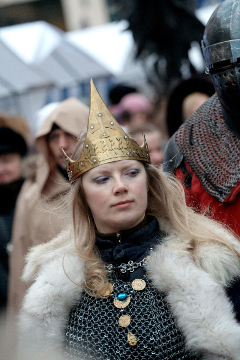A woman dressed in medieval attire stands among a crowd at the 2012 Kazjukas Festival. She wears a golden crown with intricate designs, chainmail, and a fur-trimmed cloak. Her long blonde hair falls freely, and she has a focused expression. People in similar historical costumes and modern winter wear are seen in the background.