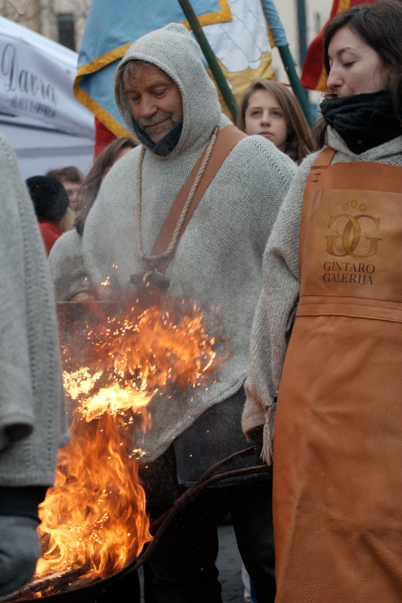 A man in a hooded garment and leather apron tends to a large fire outdoors, flames rising high. Beside him stands a woman in similar attire, "Gintaro Galerija" written on her apron. This scene, reminiscent of the 2012 Kazjukas festival, is watched by a group of hooded onlookers with colorful flags behind them.