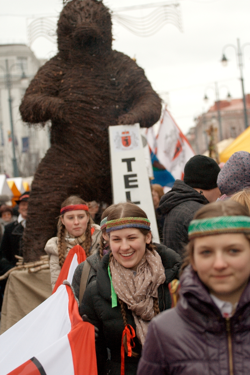 Group of people at an outdoor festival in 2012, some wearing traditional headbands. A large wicker bear sculpture is prominently displayed in the background. The people are holding flags and signs, celebrating Kazjukas. The event appears festive, with a crowd and tents visible in the background.