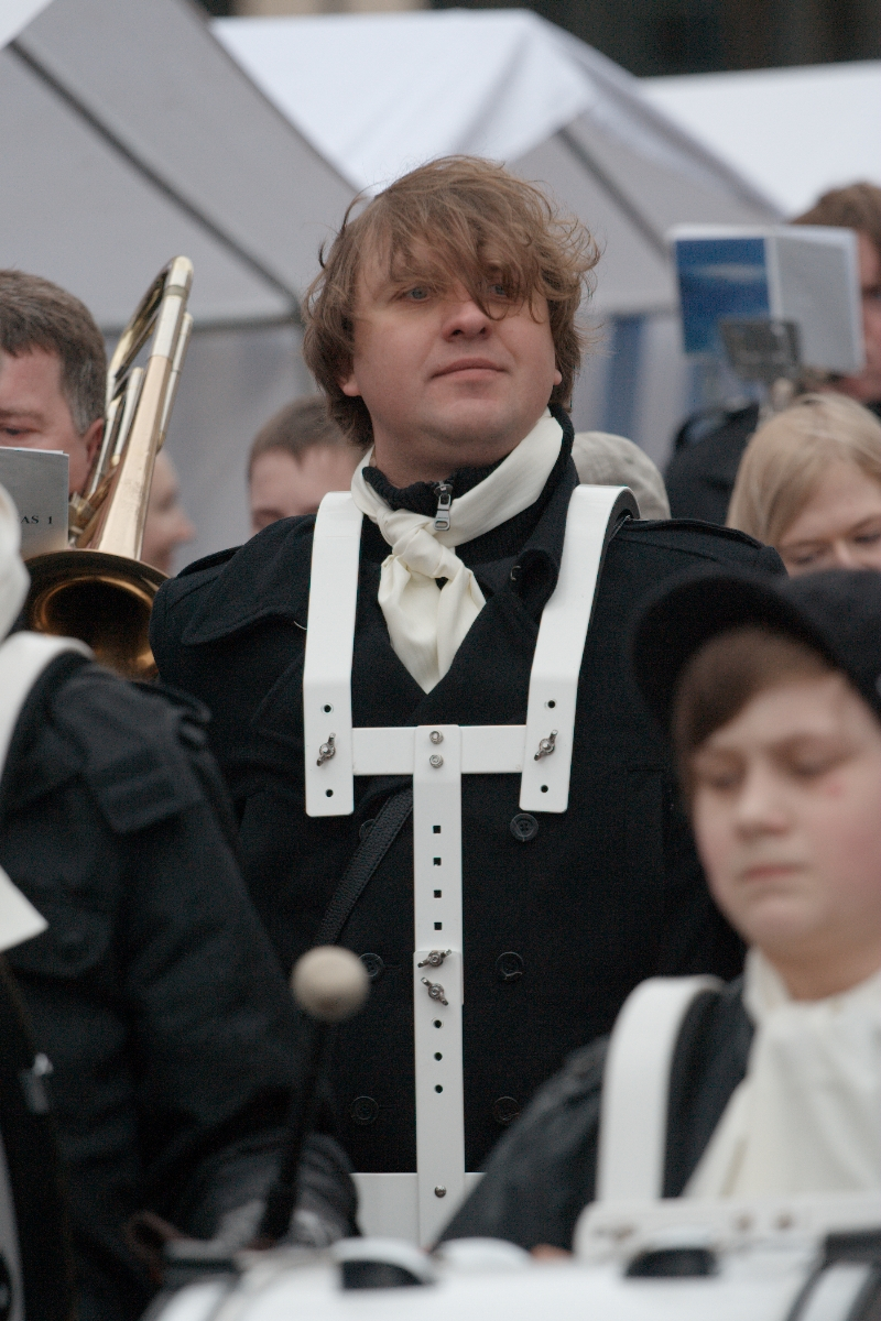 A man, wearing a black coat and a white chest strap, stands amidst a crowd at Kazjukas 2012. His brown hair is swept over one eye, and he holds a trombone. People in the background are dressed in similar attire, suggesting a formal event or parade. Another person in the foreground is out of focus.