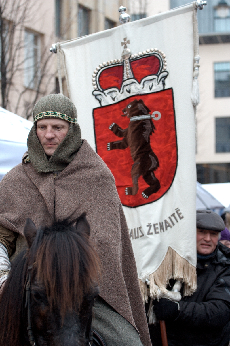 A person dressed in medieval attire, including a chainmail hood and brown cloak, rides a dark horse. Behind them is a large banner featuring a red shield with a bear standing upright, a crown, and the text "Auks Žemaitė." A modern-clothed figure stands in the background with "Kazjukas 2012" subtly visible on their shirt.