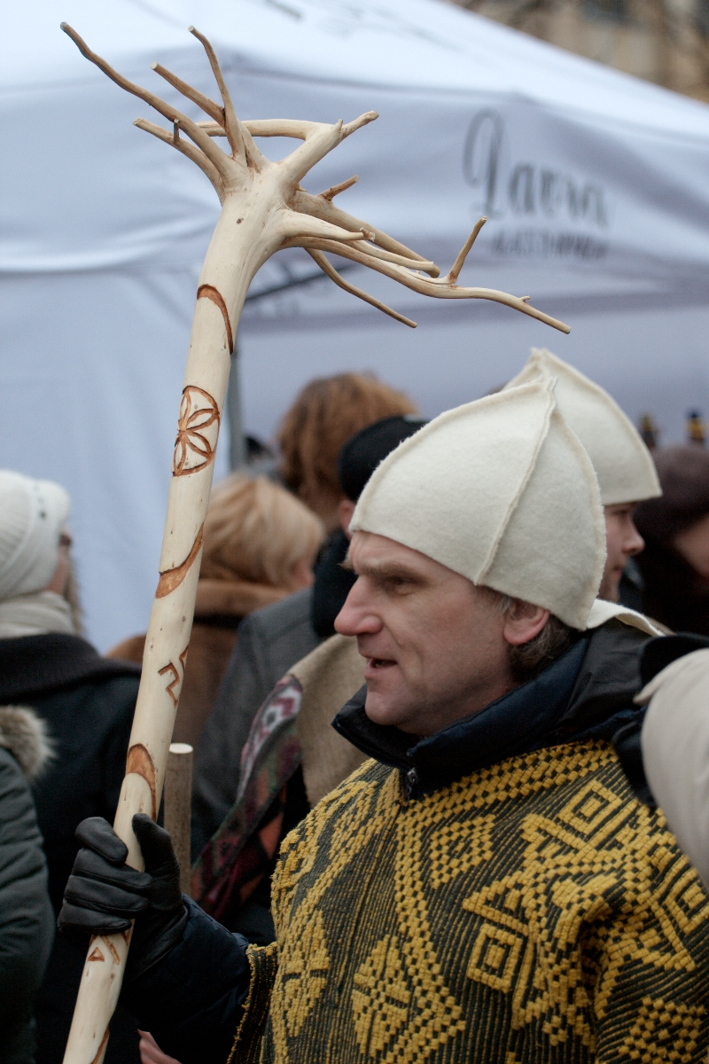 A person wearing a white felt hat and a yellow geometric-patterned garment holds a large wooden staff with intricate carvings and a tree-branch top. They are part of the Kazjukas 2012 crowd gathered in front of a white tent. The atmosphere appears to be festive, possibly during a cultural or traditional event.