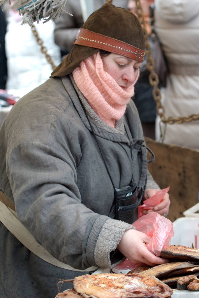 A person dressed in a heavy, old-style coat and a brown hat is working at the Kazjukas 2012 outdoor market. They have a pink scarf wrapped around their head and neck. They are handling a piece of meat, placing it in a plastic bag. The background shows a chain and other market activity.