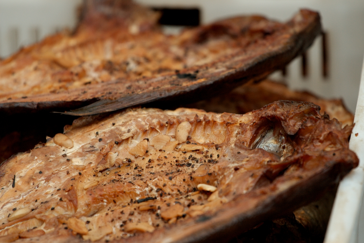 Close-up of grilled fish fillets with crispy, golden-brown skin and visible seasoning. The fish is placed in a container, displaying layers of flaky texture and scattered spices. Some bones protrude from the meat, giving it a rustic, freshly-cooked appearance reminiscent of Kazjukas 2012 standards.