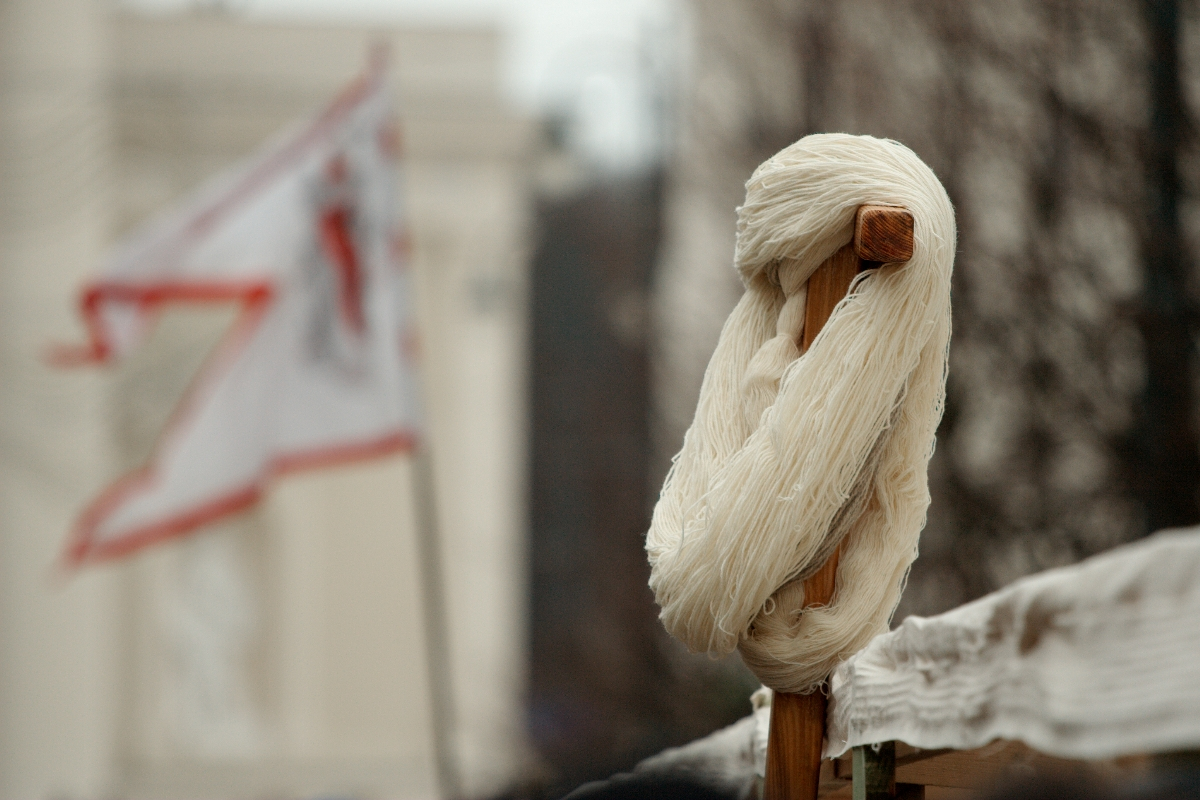 A coil of off-white yarn is wound around a small wooden post in the foreground. In the blurred background, a white flag with red markings waves, partially obscured. The setting, reminiscent of 2012, includes out-of-focus dark brown tree branches and a light-colored building, perhaps inspired by Kazjukas.
