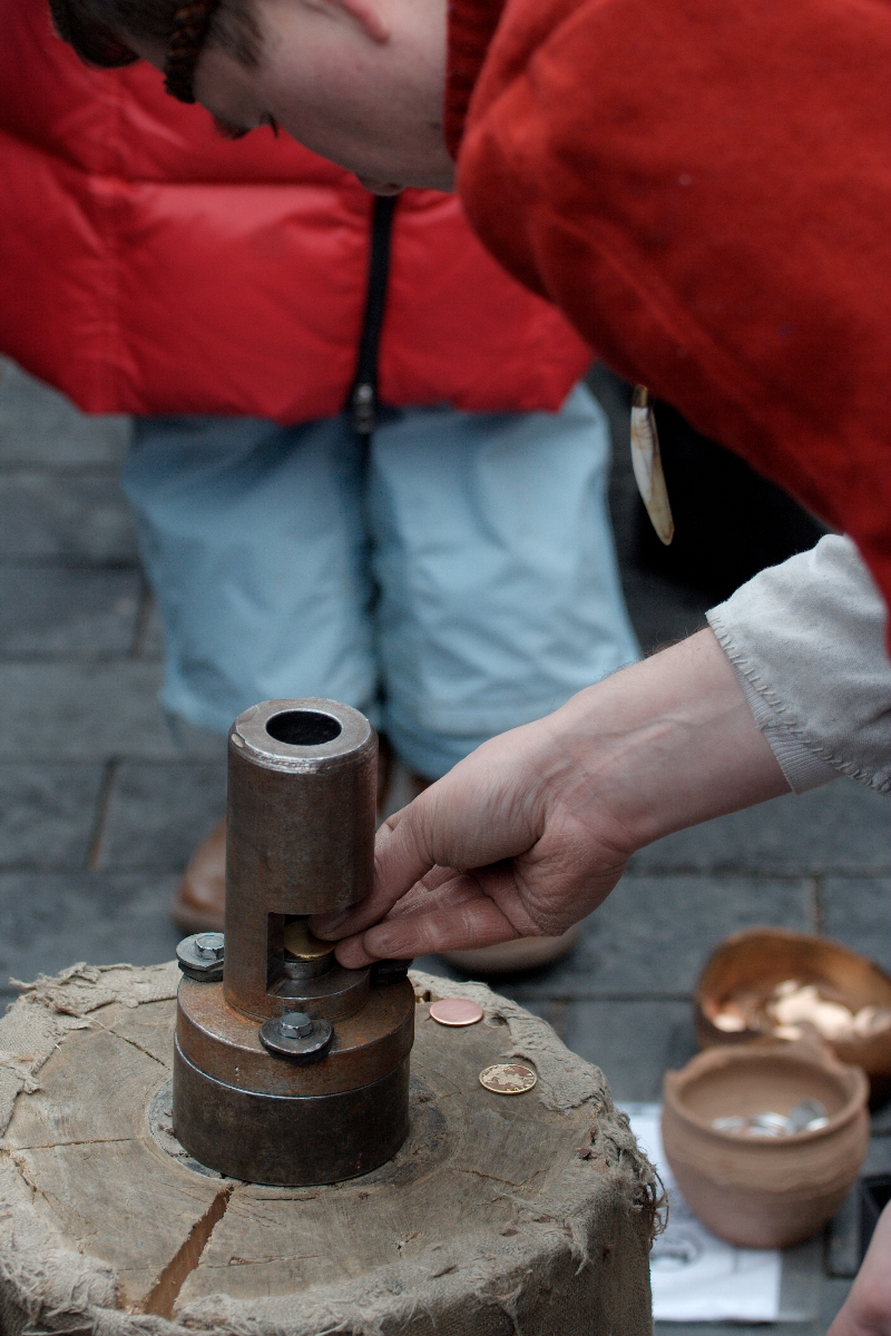 A person is using a coin-stamping device on a wooden block. The person, dressed in red and beige, carefully positions a coin for stamping. Another individual in a red coat and blue pants stands in the background. Nearby are two bowls and a paper with illustrations, reminiscent of Kazjukas 2012.