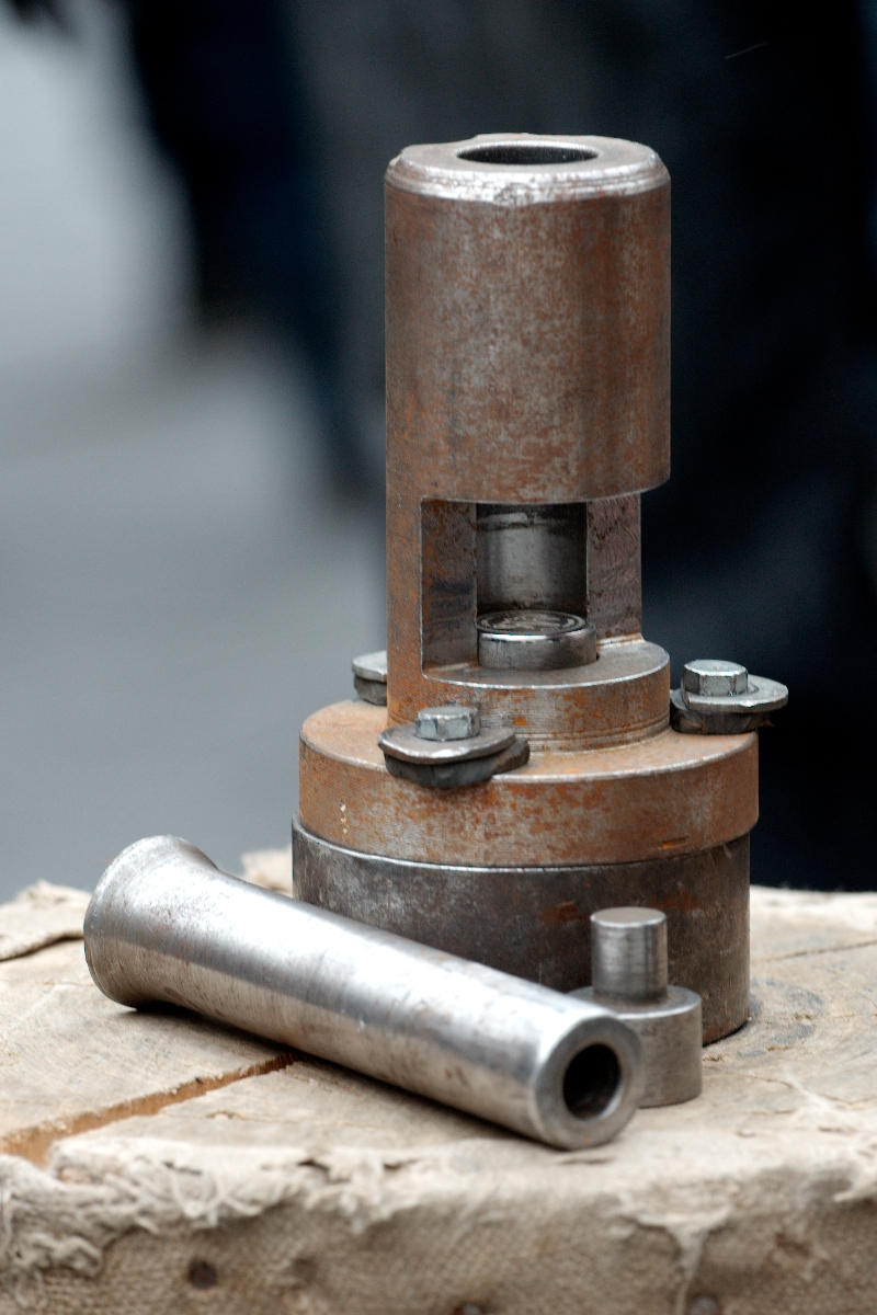 A weathered metal machine component from the Kazjukas 2012 collection, featuring bolts, a cylindrical structure, and a detachable piece—possibly a valve or industrial tool—sits on a wooden surface. The blurred background suggests that the focus is on this intricate machinery part.